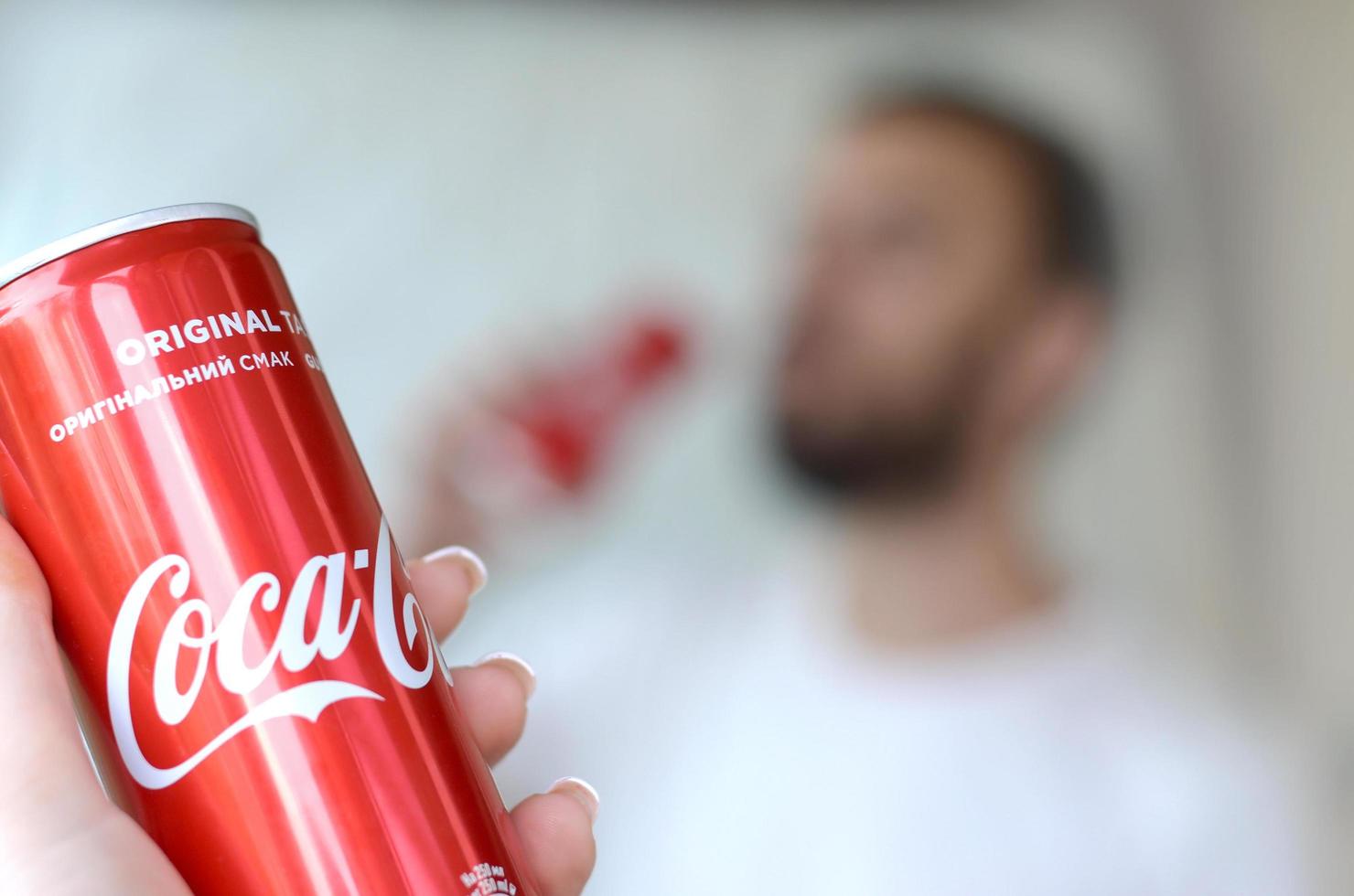 KHARKOV. UKRAINE - MAY 2, 2019 Caucasian man drinks Coca-Cola drink in garage interior and male hand presents one Coca cola red can in focus photo