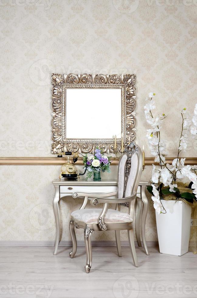 A fragment of a retro interior with a chair and table, on which is a telephone and a vase of flowers. Above the table hangs an empty picture frame for your photo