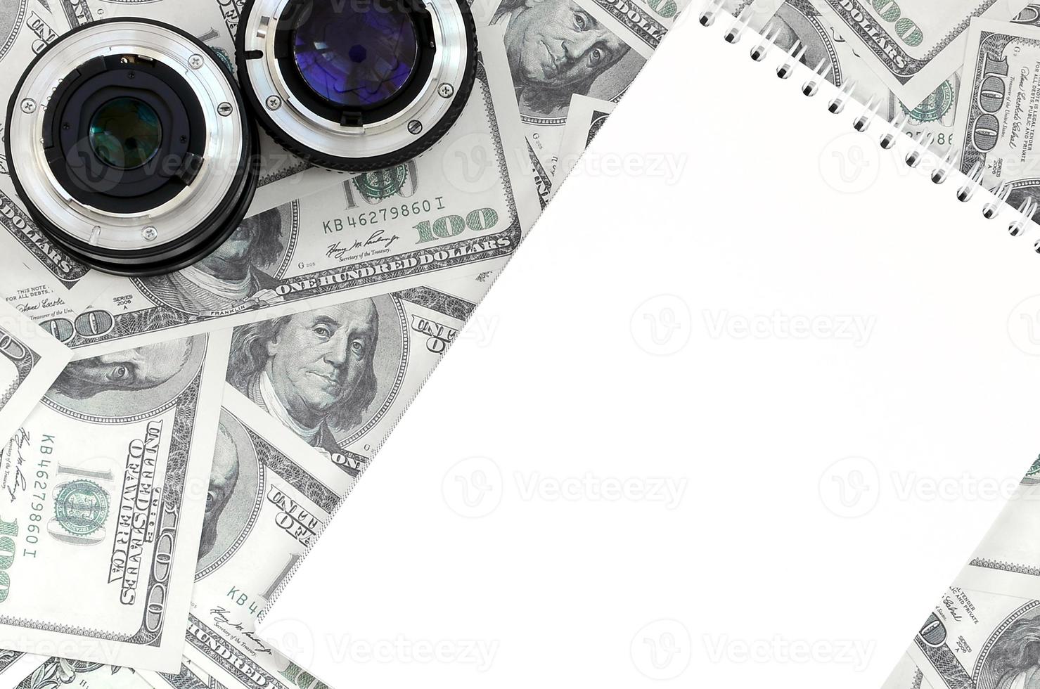 Two photographic lenses and white notebook lie on the background of a lot of dollar bills. Space for text photo