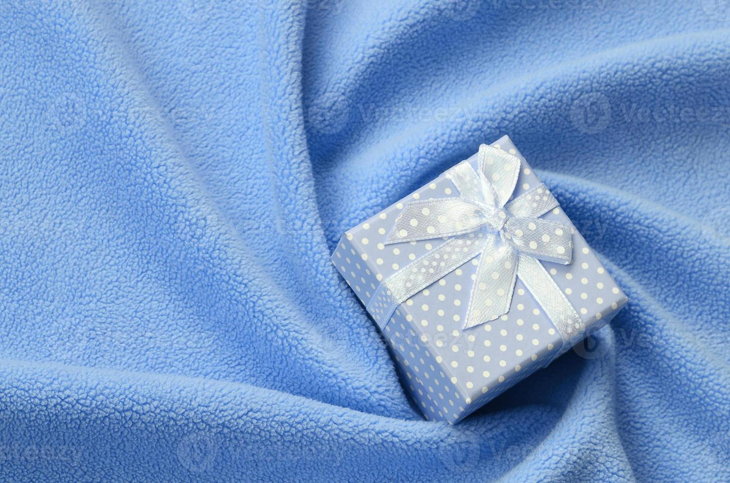 A small gift box in blue with a small bow lies on a blanket of soft and furry light blue fleece fabric with a lot of relief folds. Packing for a gift to your lovely girlfriend photo