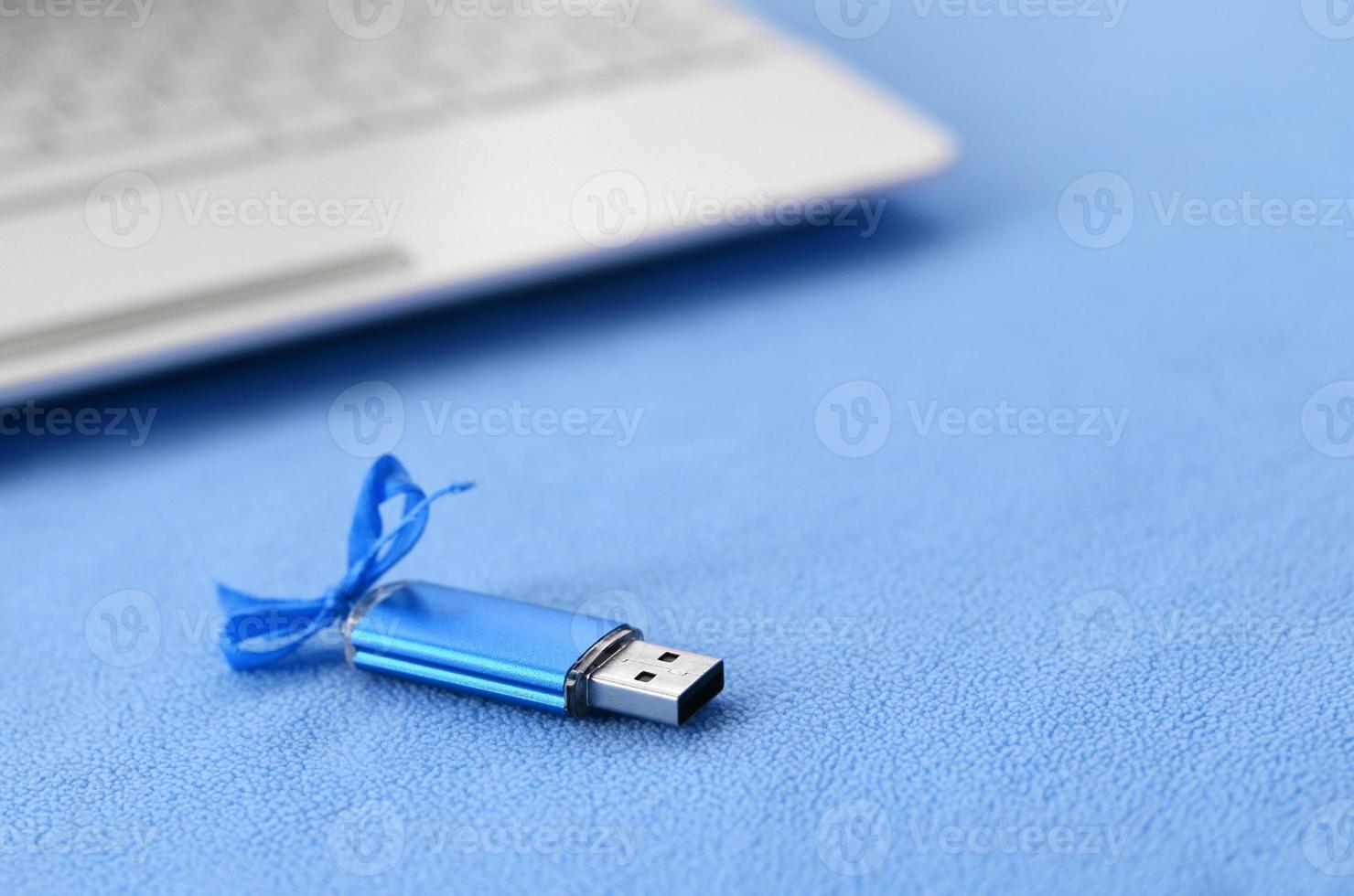 Brilliant blue usb flash memory card with a blue bow lies on a blanket of soft and furry light blue fleece fabric beside to a white laptop. Classic female gift design for a memory card photo