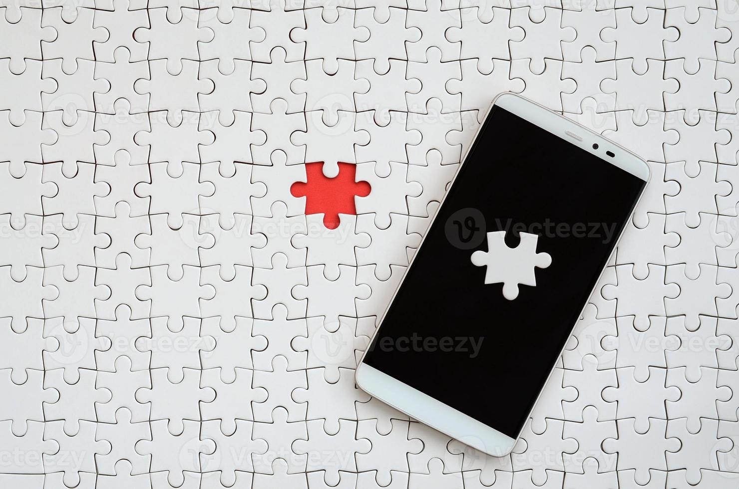 A modern big smartphone with several puzzle elements on the touch screen lies on a white jigsaw puzzle in an assembled state with missing elements photo