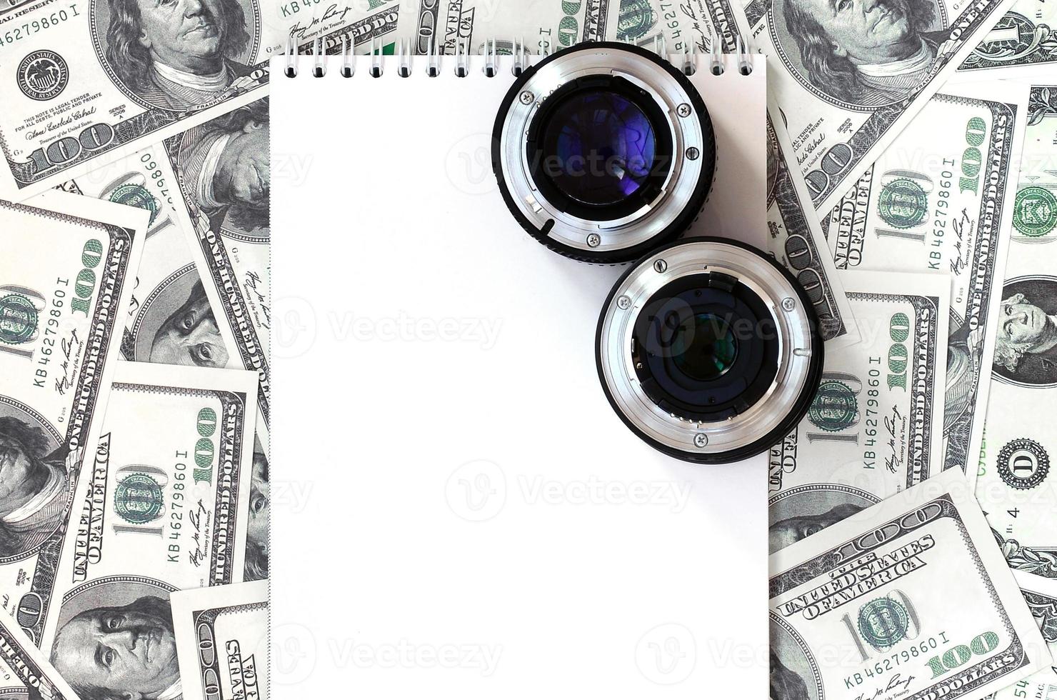 Two photographic lenses and white notebook lie on the background of a lot of dollar bills. Space for text photo
