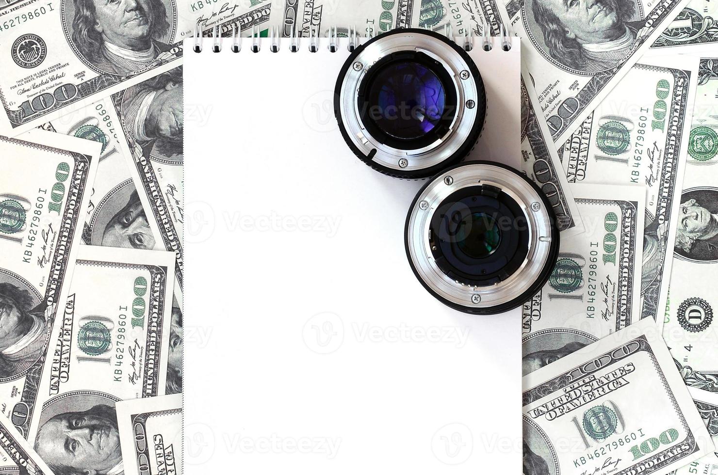 dos lentes fotográficos y un cuaderno blanco se encuentran en el fondo de muchos billetes de dólar. espacio para texto foto