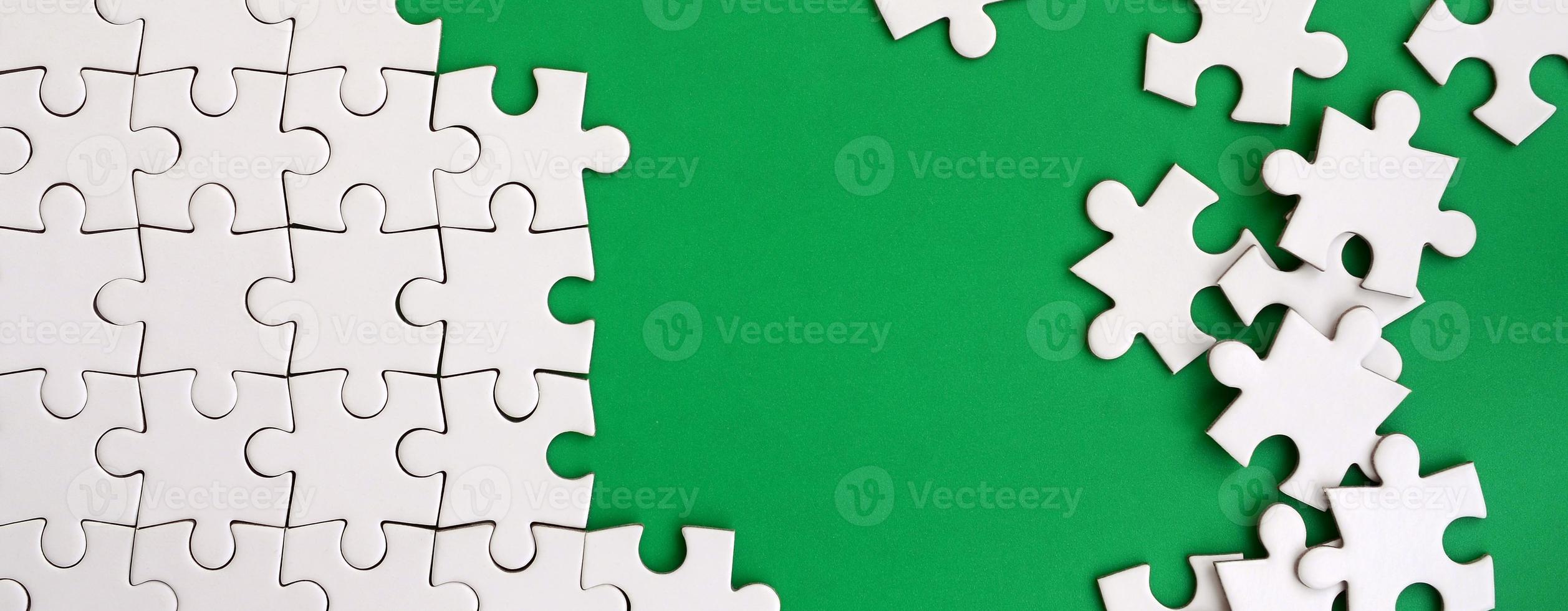 Fragment of a folded white jigsaw puzzle and a pile of uncombed puzzle elements against the background of a green surface. Texture photo with space for text