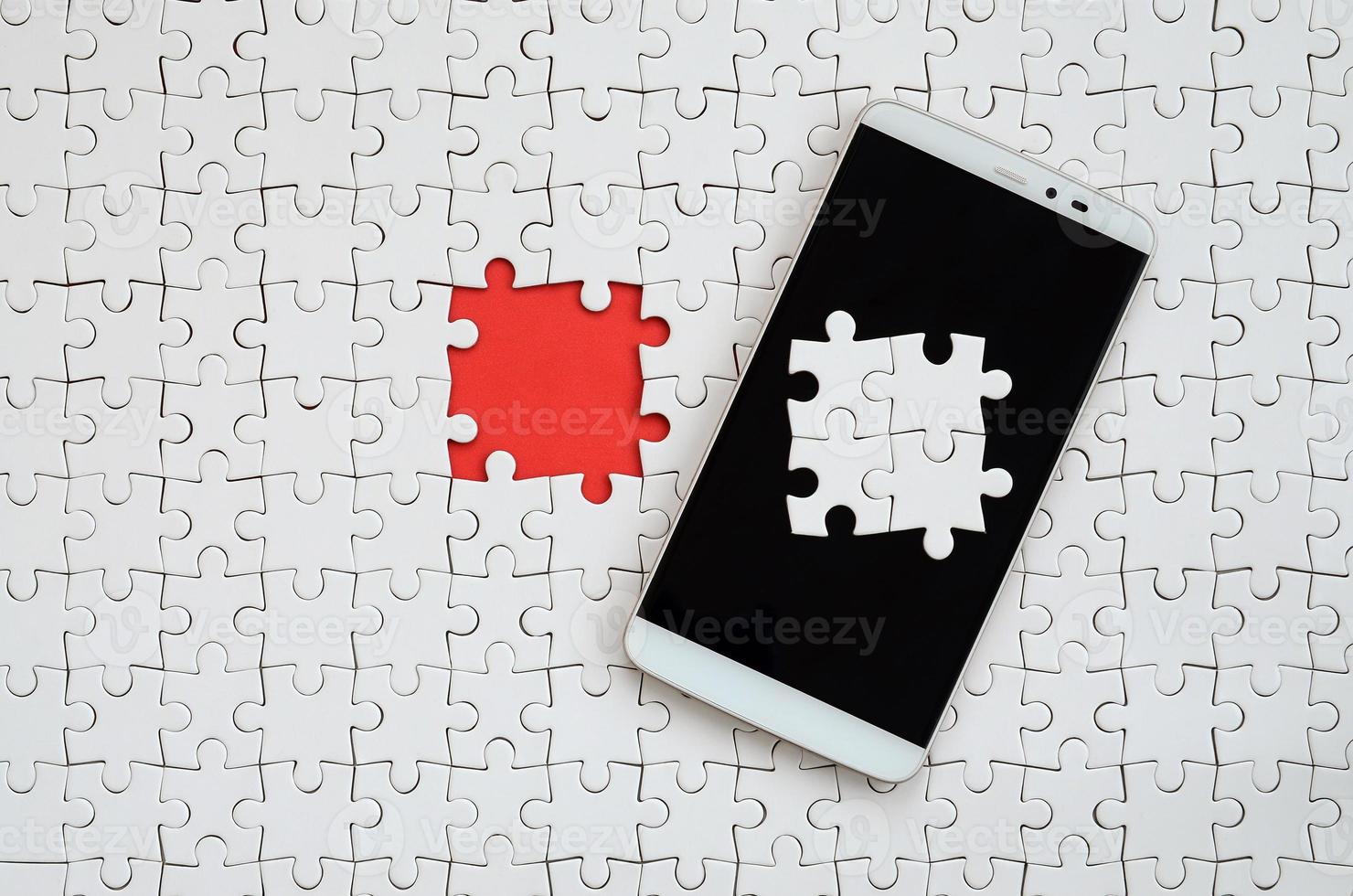 A modern big smartphone with several puzzle elements on the touch screen lies on a white jigsaw puzzle in an assembled state with missing elements photo