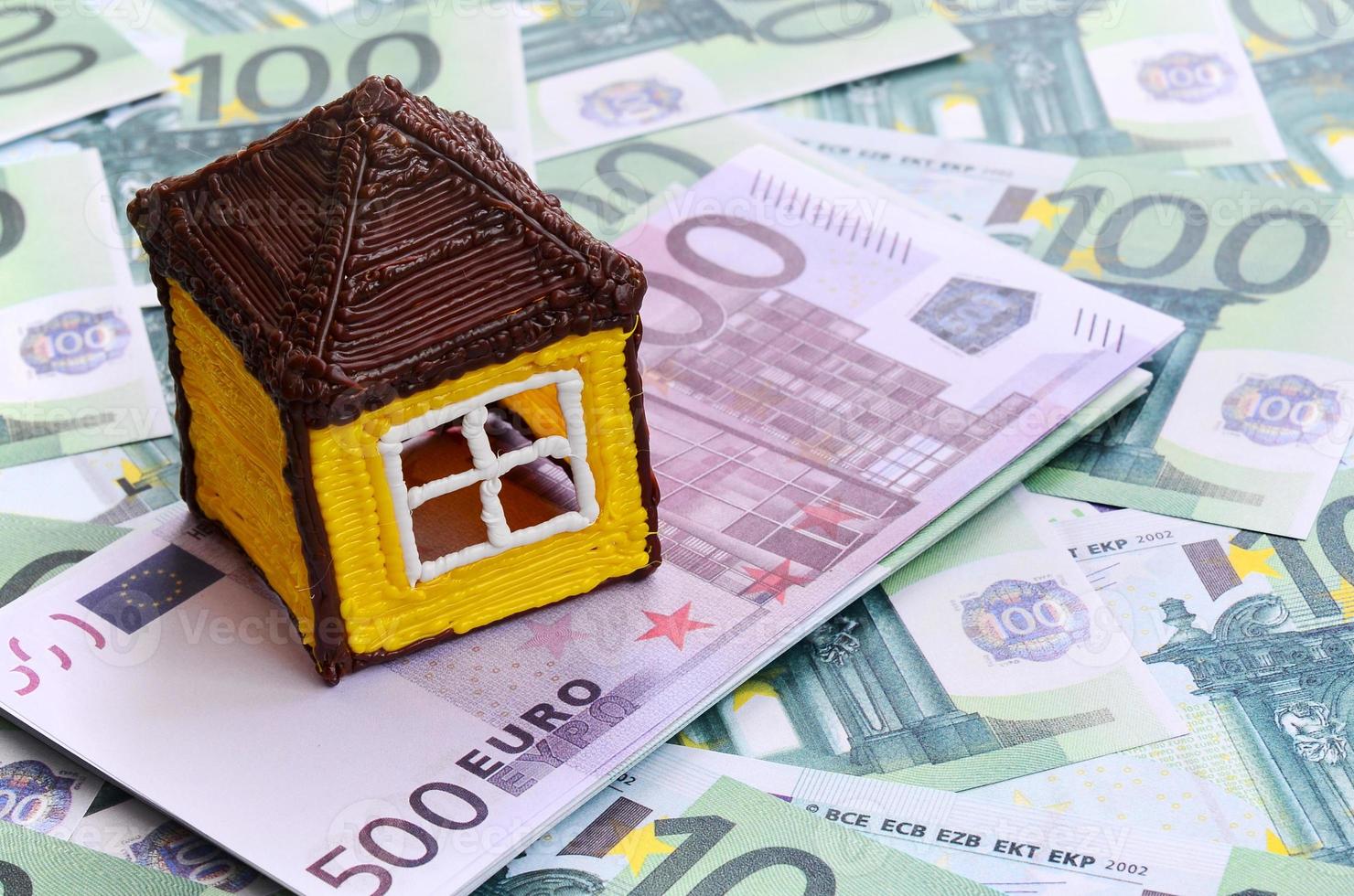 Small toy house is lies on a set of green monetary denominations of 100 euros. A lot of money forms an infinite heap photo