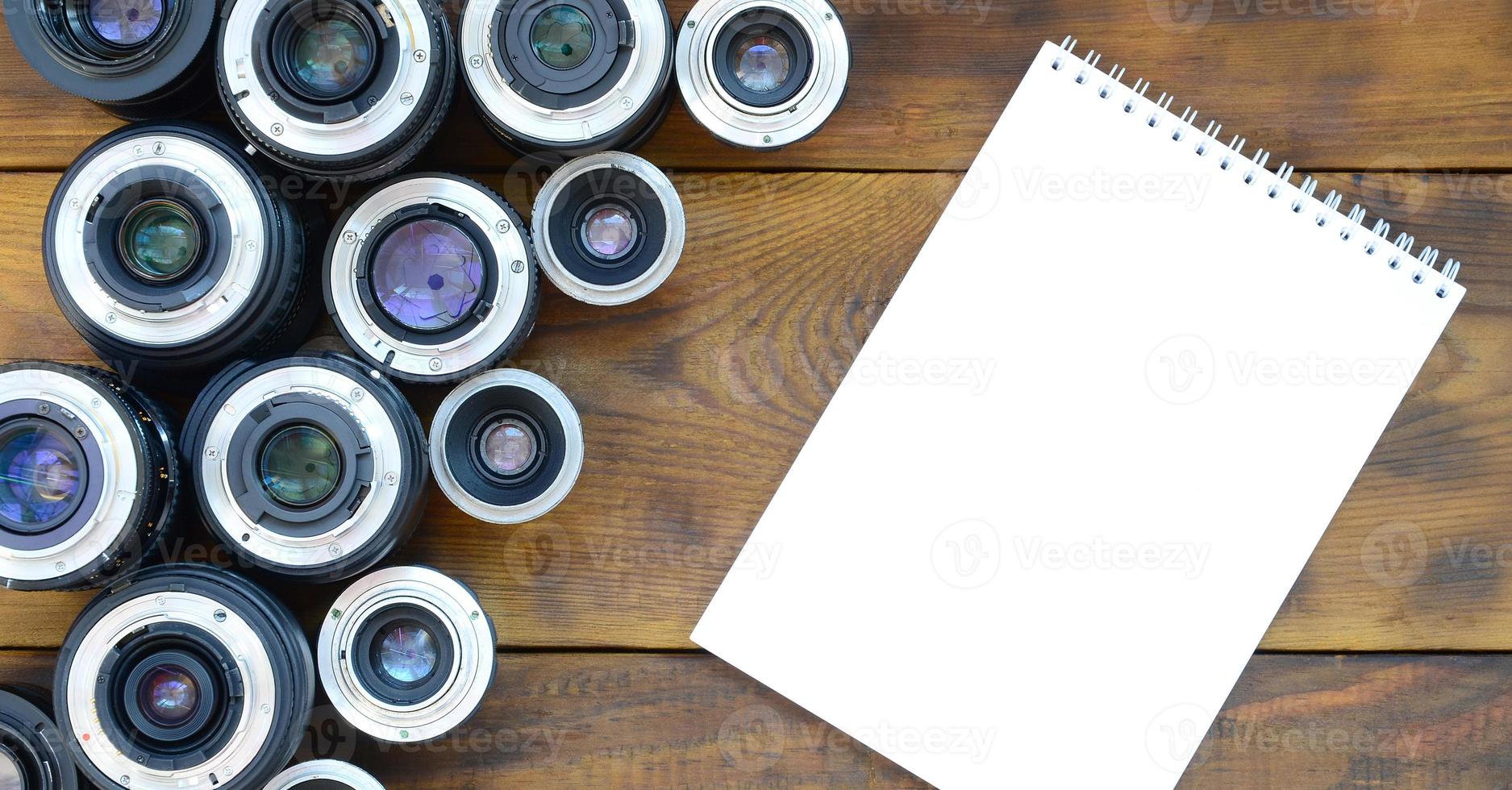 Several photographic lenses and white notebook lie on a brown wooden background. Space for text photo