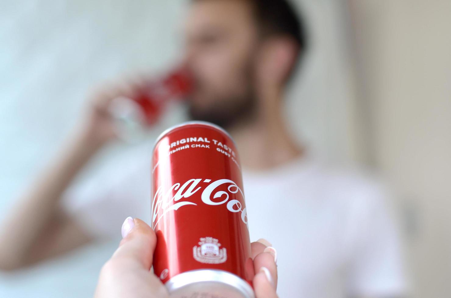 KHARKOV. UKRAINE - MAY 2, 2019 Caucasian man drinks Coca-Cola drink in garage interior and male hand presents one Coca cola red can in focus photo