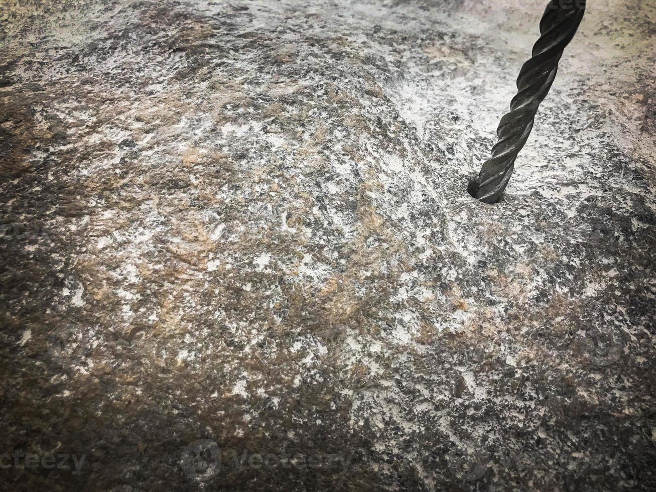 A sturdy, hard metal iron drill bit drills a hole in a large gray stone. Close view. The background photo