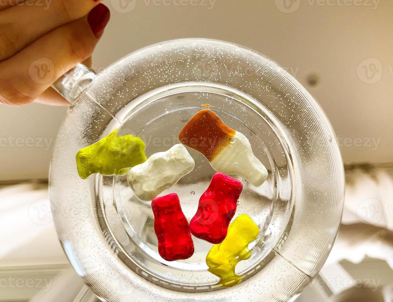 transparent mug, round and large, filled with water. at the bottom of the mug are gummy bears and bottles of lemonade. girl holding a mug in her hands. swimming pool for gelatin candy photo