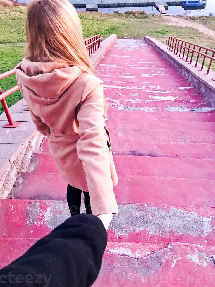 A beautiful girl in a brown coat with long blond hair leads a guy by the hand along the red stairs down to the river, lake, sea, water photo