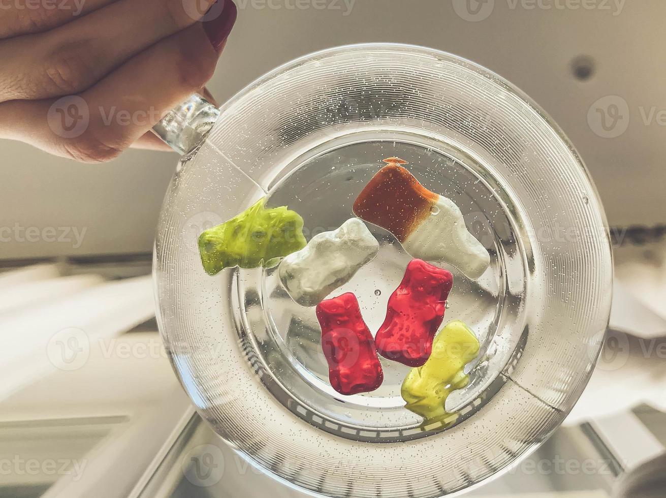 transparent mug, round and large, filled with water. at the bottom of the mug are gummy bears and bottles of lemonade. girl holding a mug in her hands. on white matte background photo