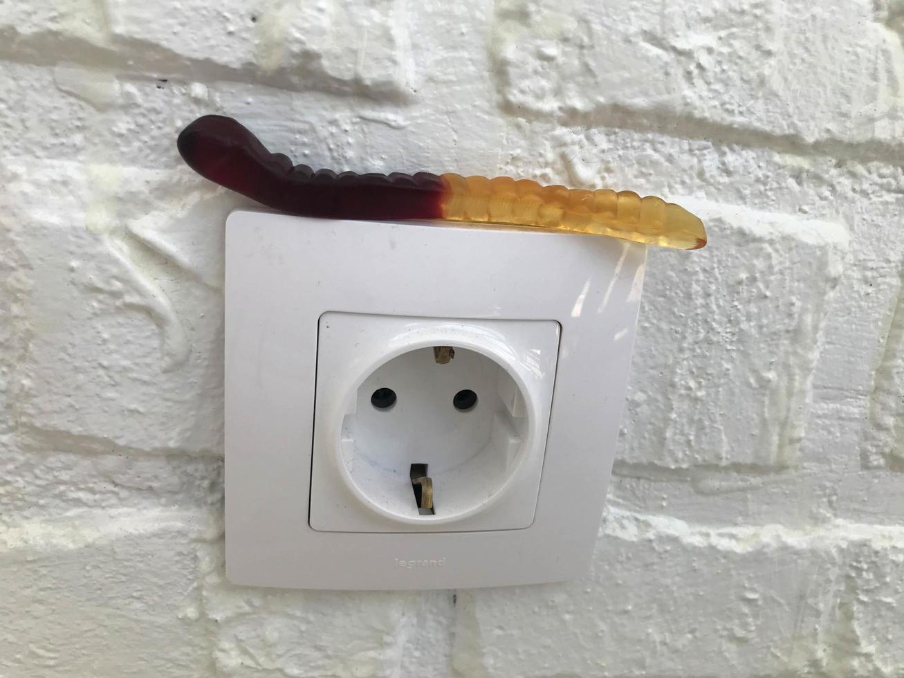 gummy worm on the background of a white brick plastered wall. a gelatinous worm lies on a white socket for electricity. delicious and sweet desserts, natural worm, halloween home decor photo