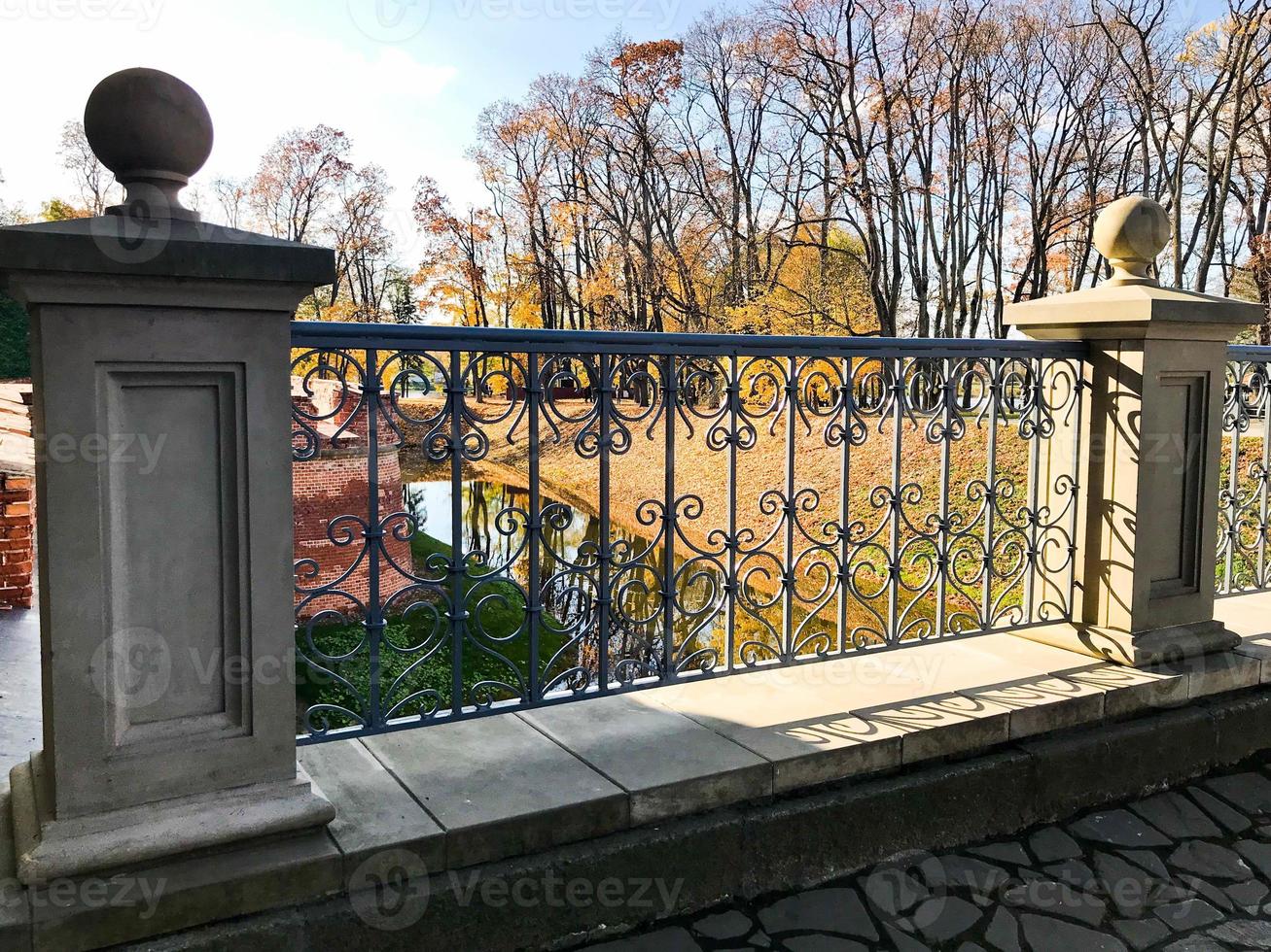 Old ancient vintage beautiful stone wrought iron railing of a bridge with patterns on the background of the river and trees with yellow leaves photo
