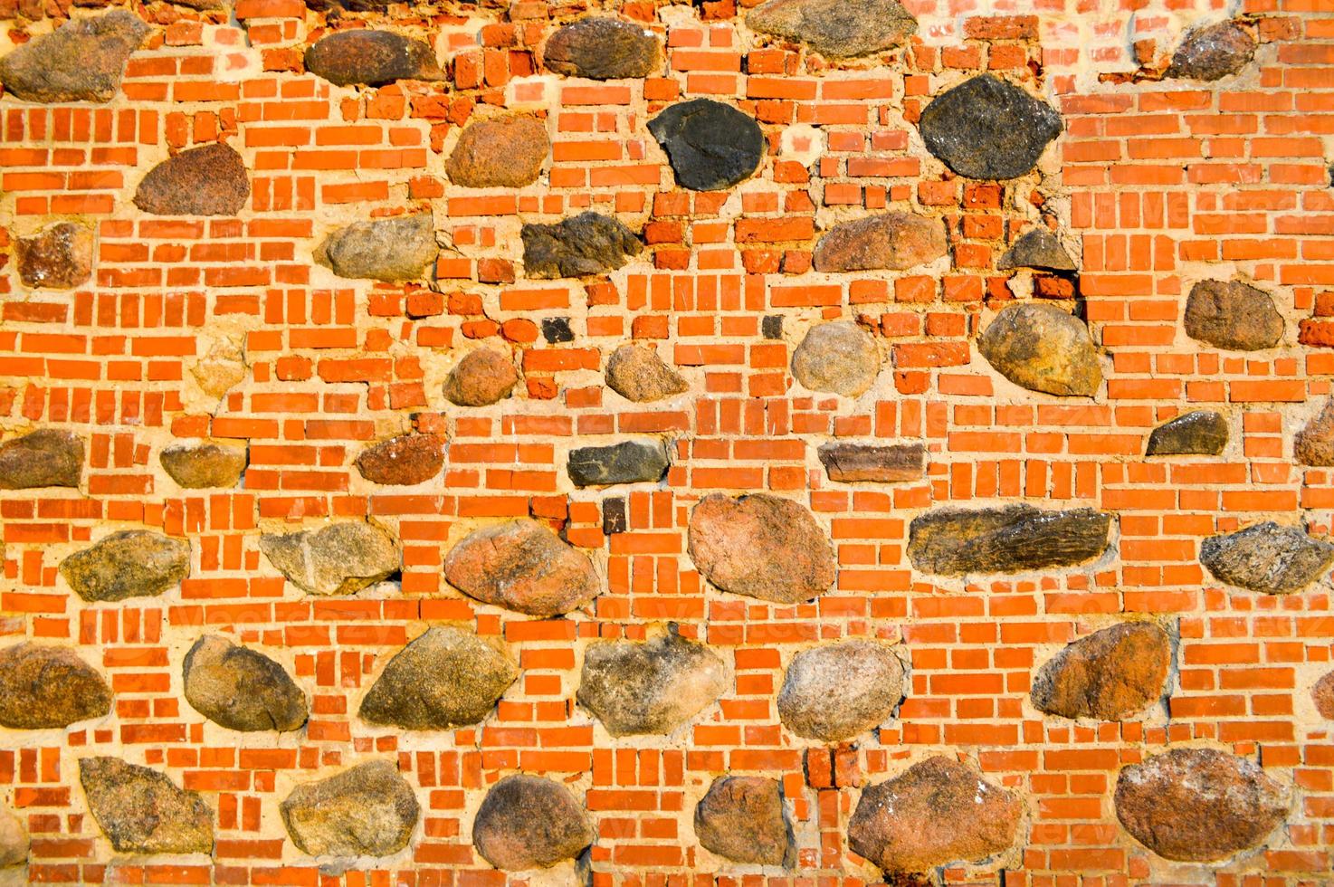 The texture of the old ancient medieval antique stone hard peeling cracked brick wall of rectangular red clay bricks and large stones, cobblestones. The background photo