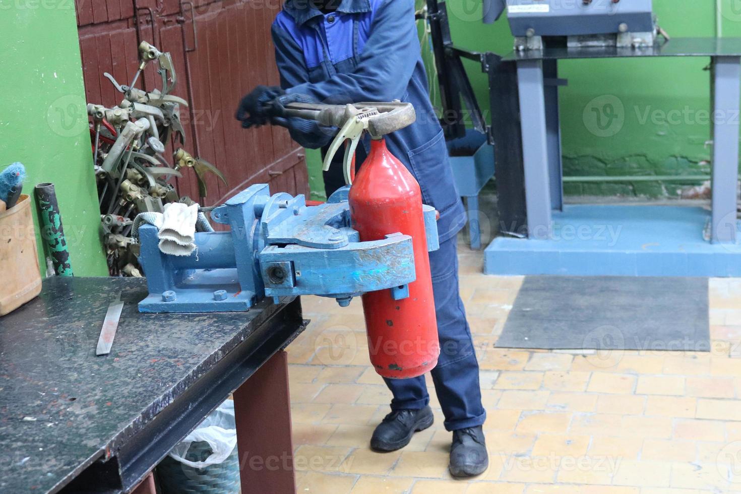 A man working on a large metal industrial vice with a huge gas wrench unscrews a nut on a red fire extinguisher cylinder in a factory workshop photo