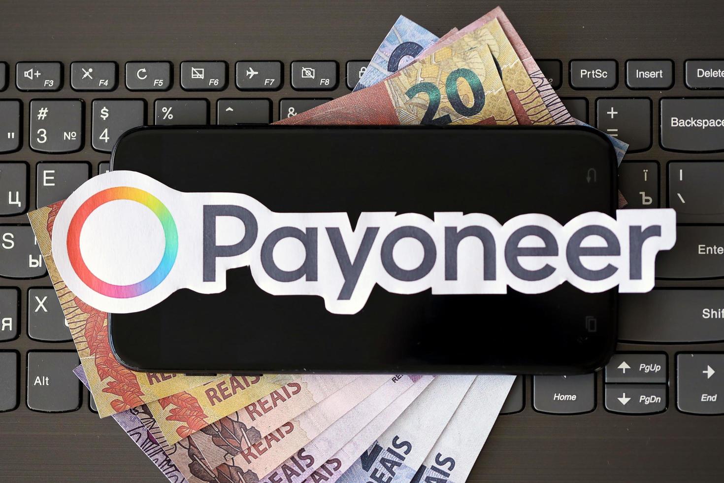 TERNOPIL, UKRAINE - SEPTEMBER 6, 2022 Payoneer paper logotype lies on black laptop with brazilian reals bills. Payoneer is American financial services company provides online money transfer photo