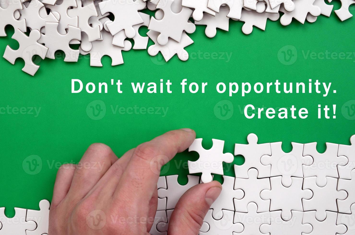 Don't wait for opportunity. Create it. The hand folds a white jigsaw puzzle and a pile of uncombed puzzle pieces lies against the background of the green surface photo