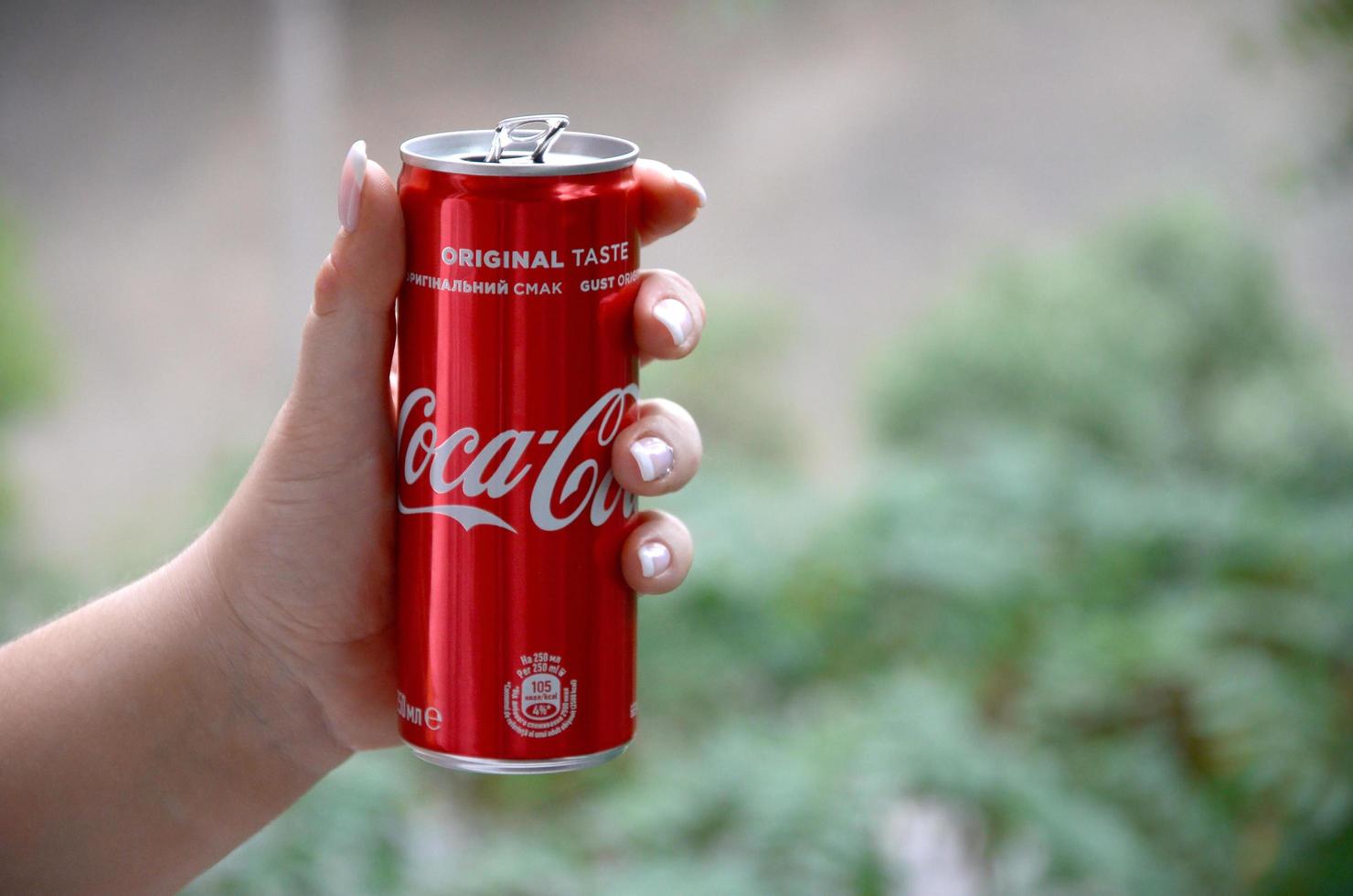 KHARKOV. UKRAINE - MAY 2, 2019 Caucasian woman holds red Coca-Cola tin can with green garden background photo