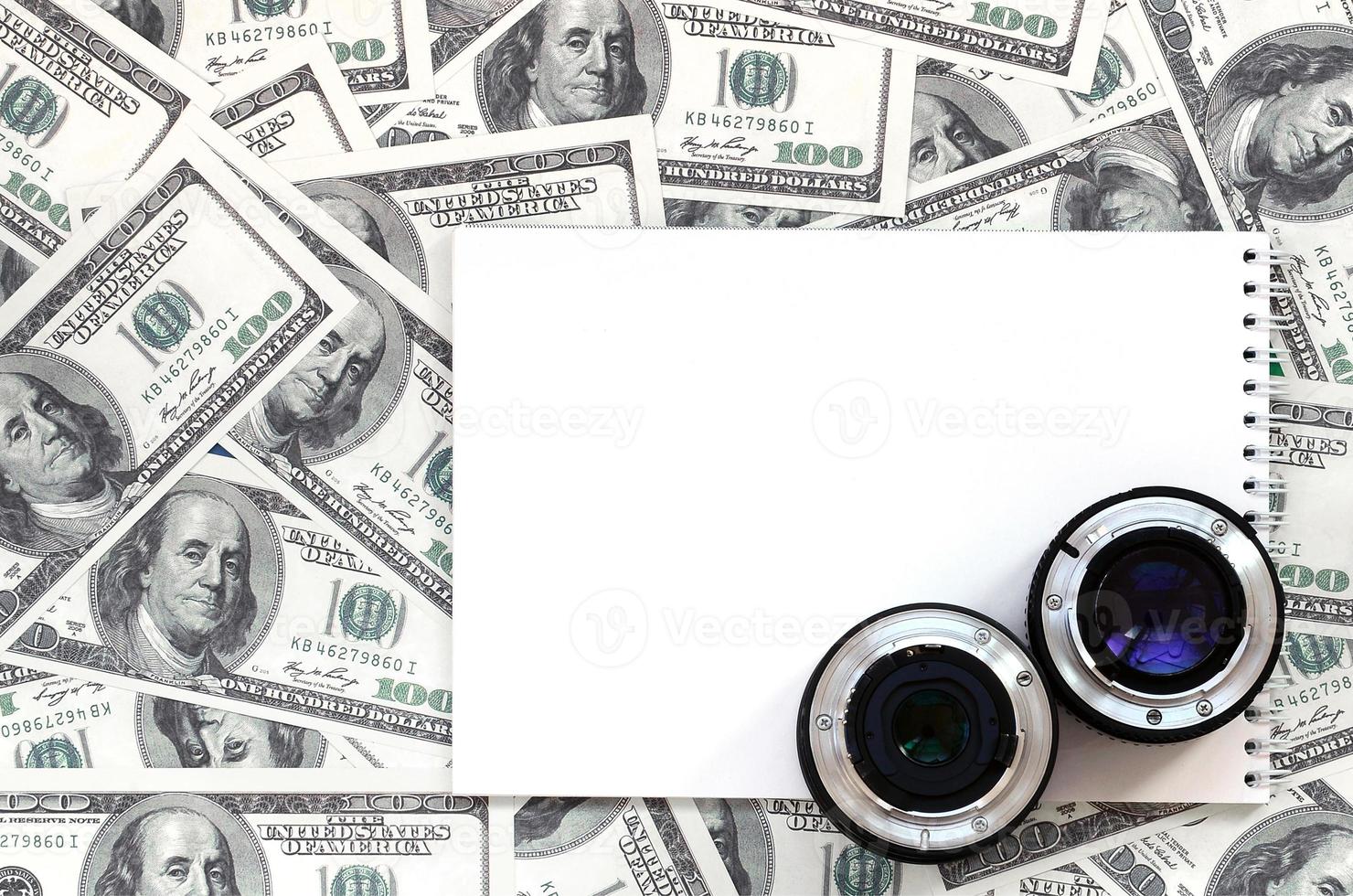 Two photographic lenses and white notebook lie on the background of a lot of dollar bills. Space for text photo