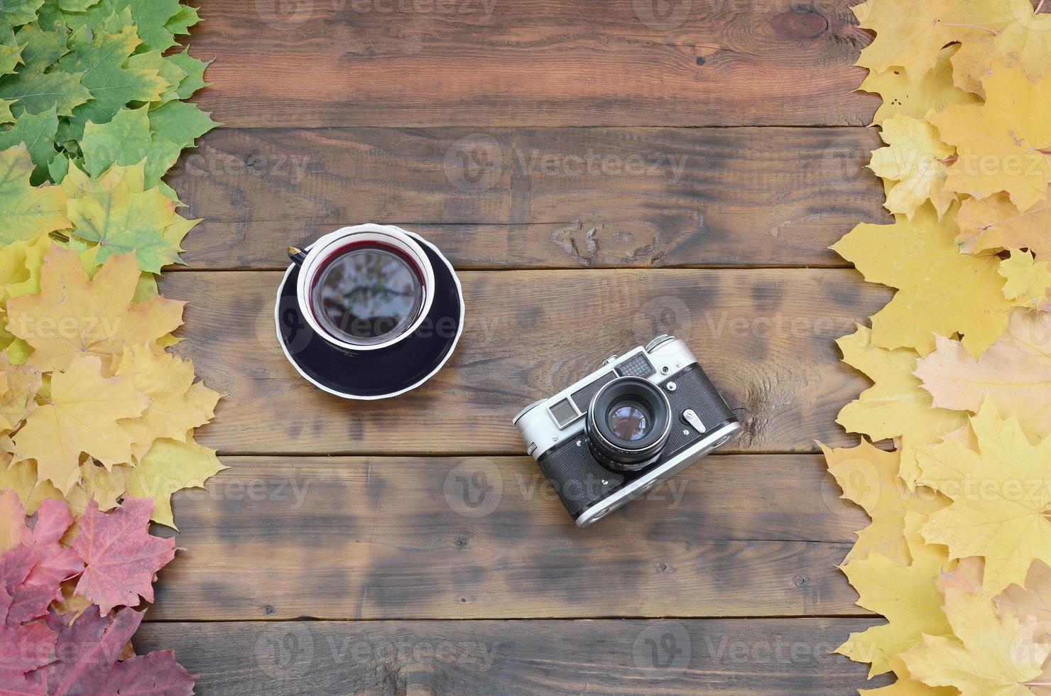 A cup of tea and an old camera among a set of yellowing fallen autumn leaves on a background surface of natural wooden boards of dark brown color photo