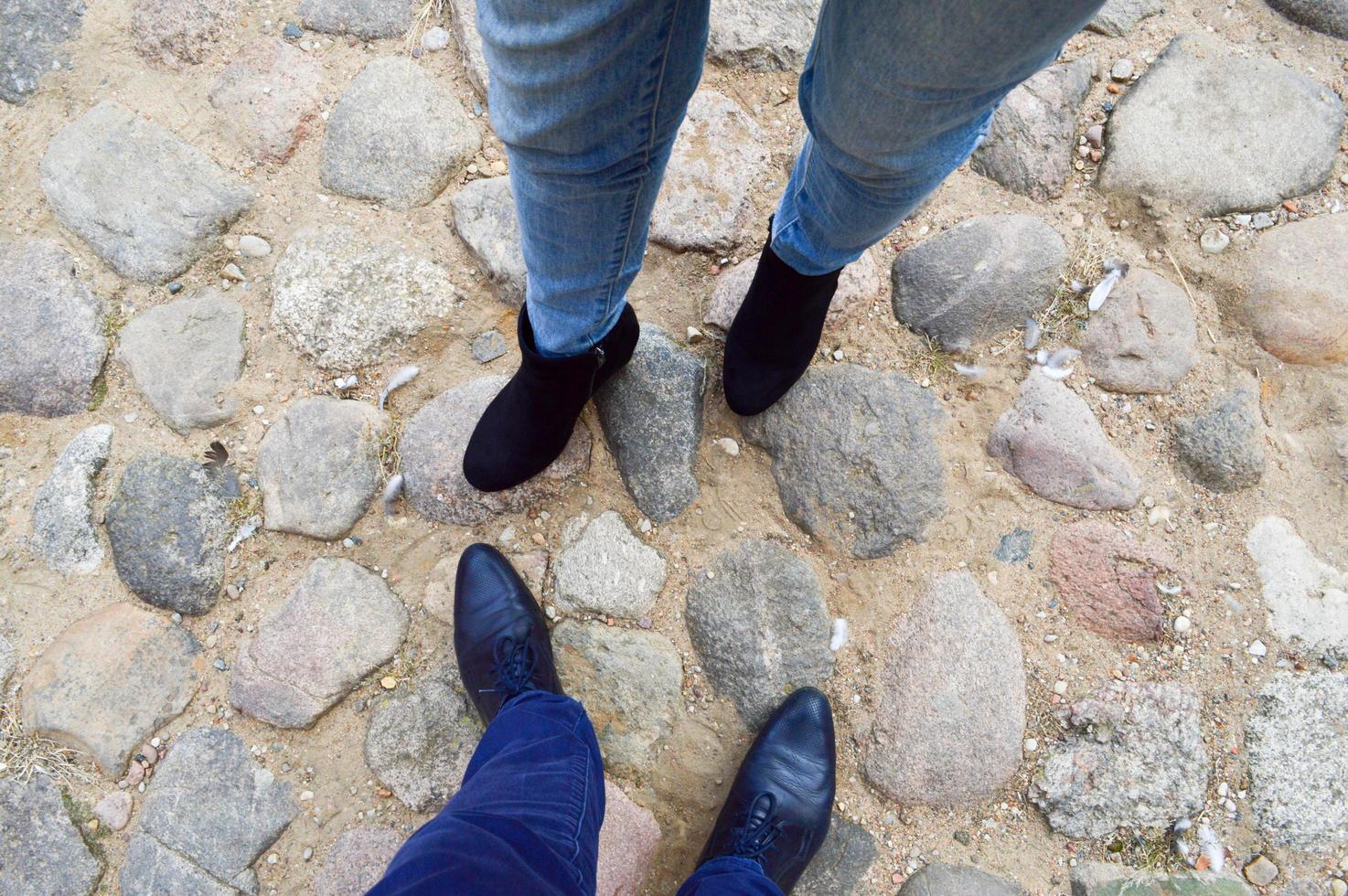 Male and female legs in leather shoes, boots on a stone road of large cobble stones opposite each other. The background photo
