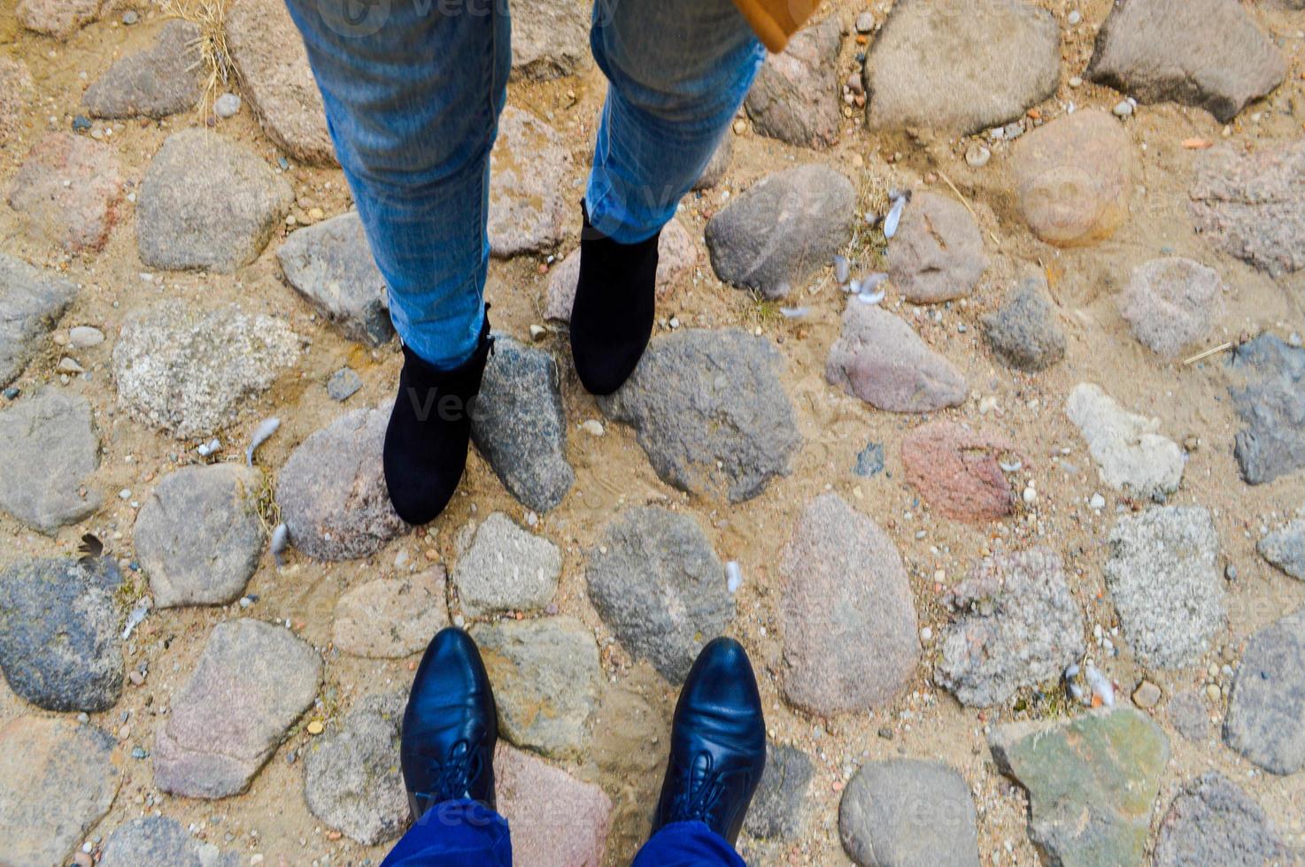 Male and female legs in leather shoes, boots on a stone road of large cobble stones opposite each other. The background photo