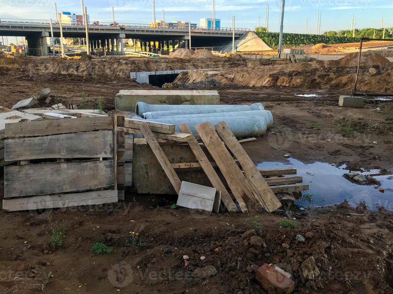 construction site, sand and mud puddles. there are wooden structures on the site. against the background of the bridge, road junction. pipes and building materials lie on the ground photo