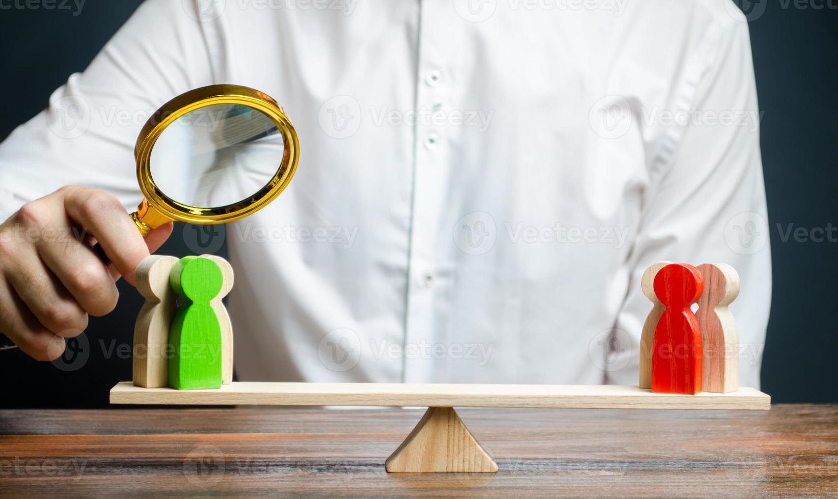 A man with a magnifying glass is looking at the rival red and green figures groups on scales. conflict resolution and the search for a compromise in the dispute. Research argument of each side photo
