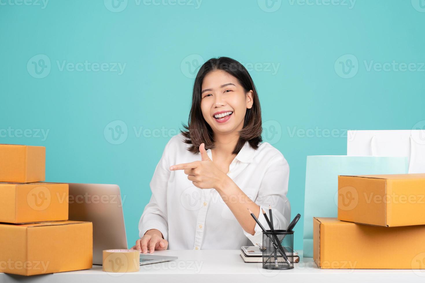 Young Asian woman in casual shirt sit work at desk with a brown cardboard box and point index finger on pc laptop. She using internet online isolated on green background. photo