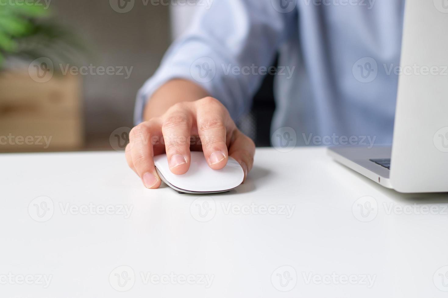 Close up of a young business man using a laptop computer with a mouse on a white desk to work at home photo