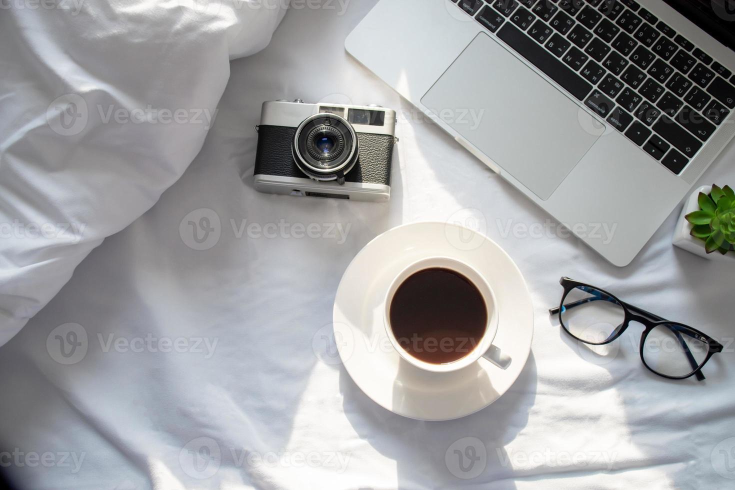 Top view, laptop computer, hot coffee, film camera and eyeglasses on the bed with morning sun. photo