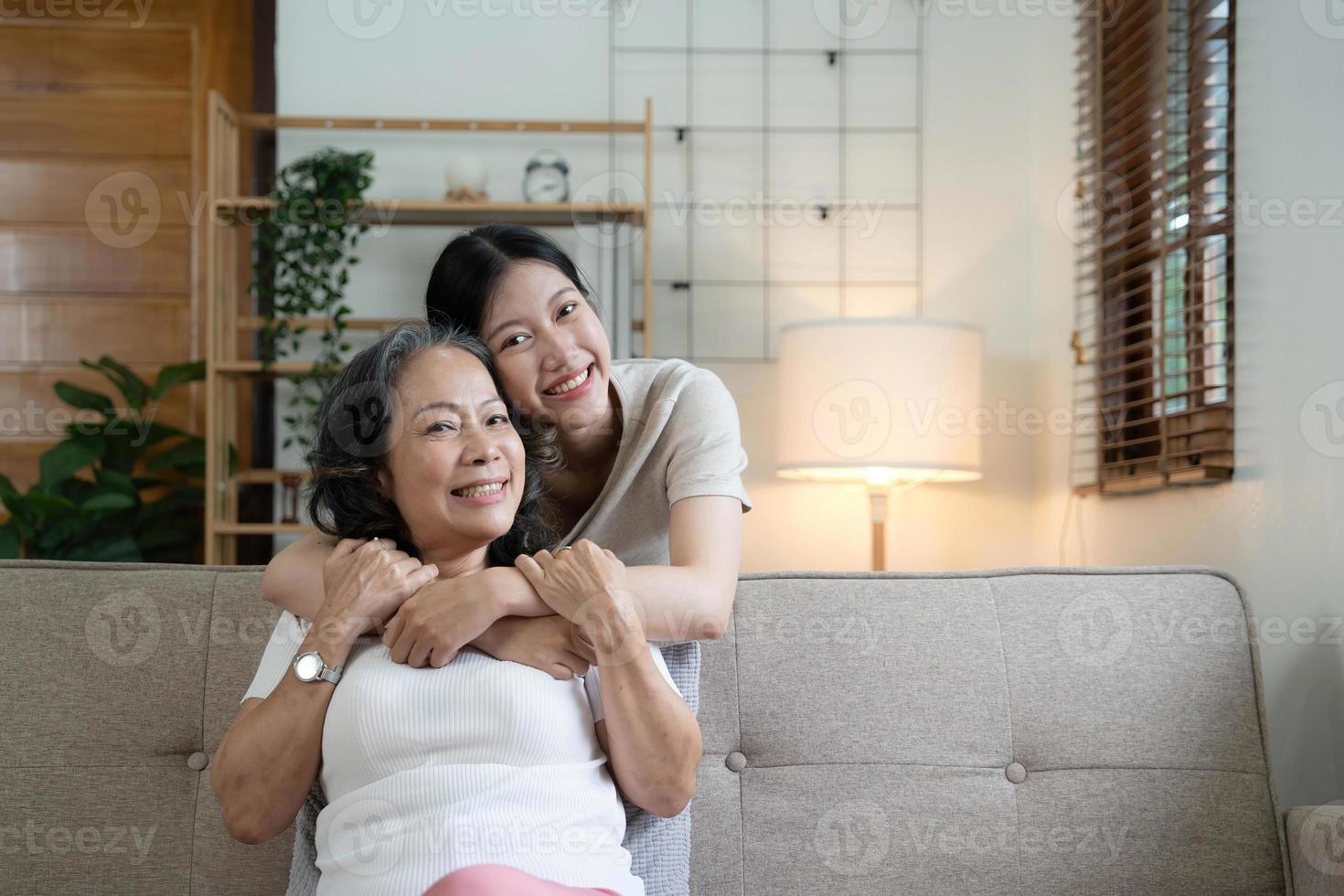 Portrait happy adult daughter and older mother hugging and holding hands, sitting on couch at home, young woman and mature mum or grandmother looking at camera, two generations family photo