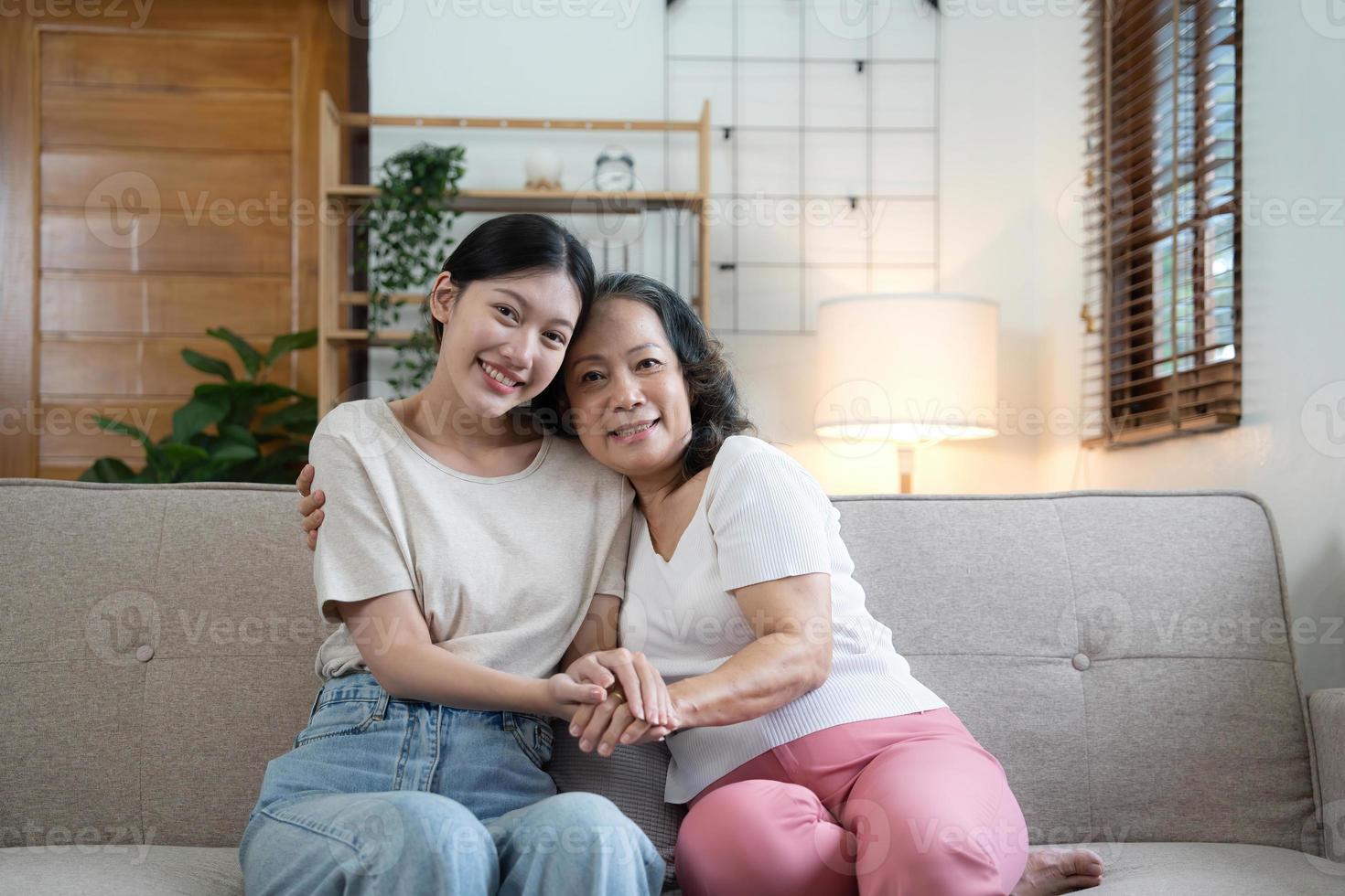 retrato feliz hija adulta y madre mayor abrazándose y tomándose de la mano, sentada en el sofá en casa, mujer joven y madre madura o abuela mirando la cámara, foto familiar de dos generaciones