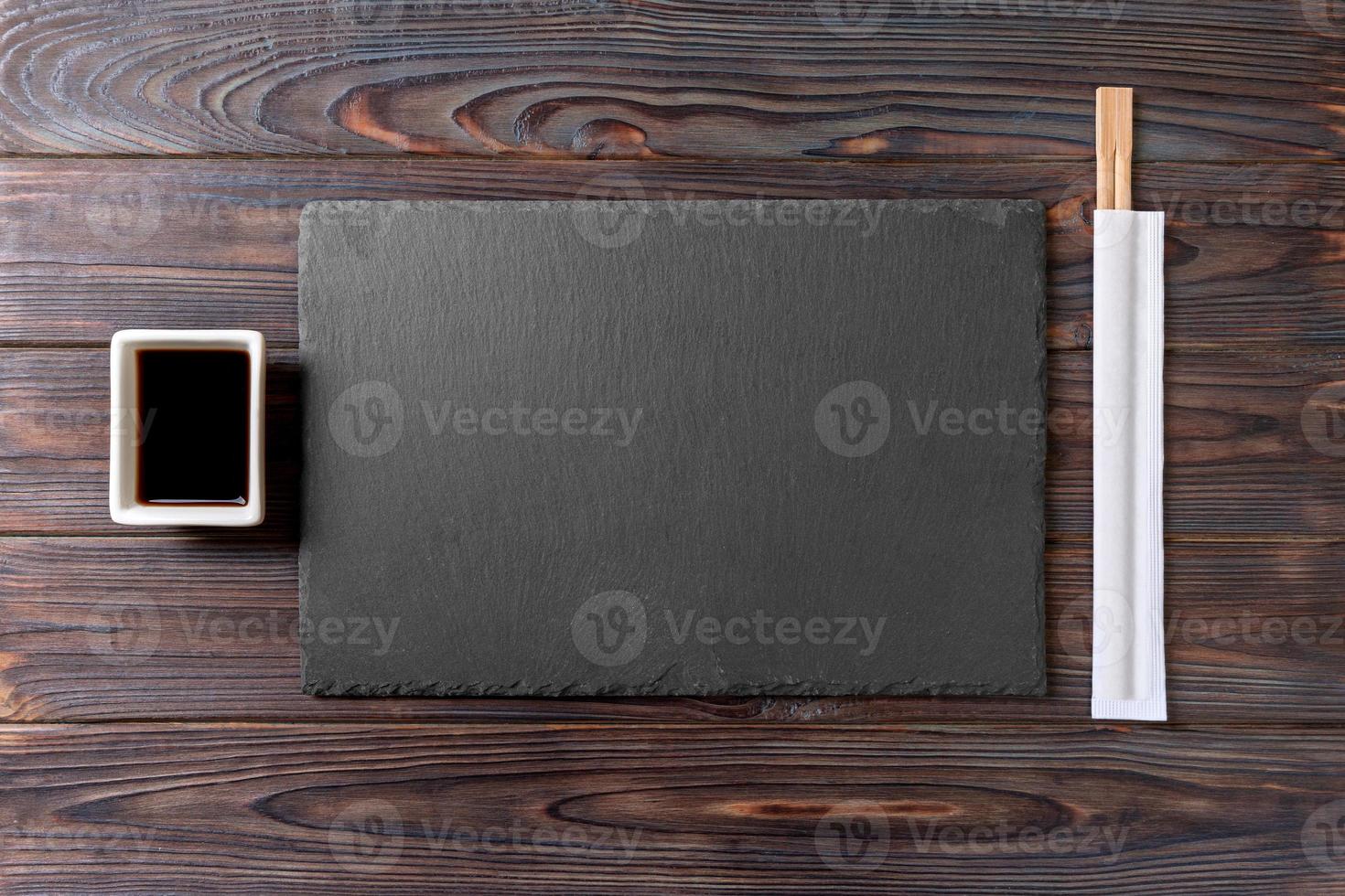 Empty rectangular black slate plate with chopsticks for sushi and soy sauce on wooden background. Top view with copy space photo