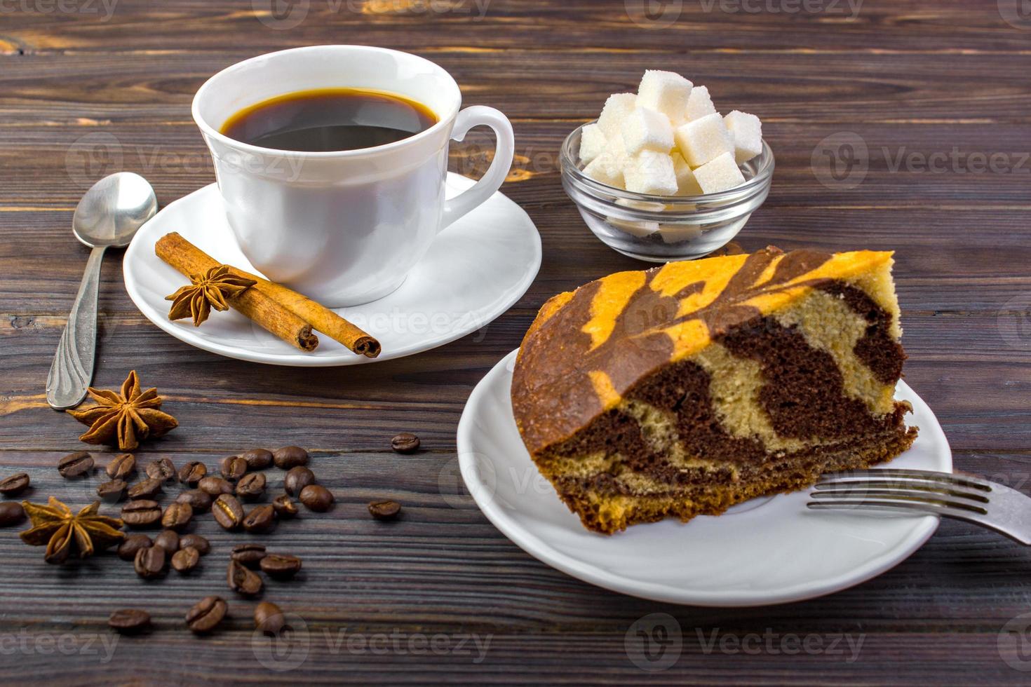 A chocolate cake on a white plate with a fork and a cup of black tea. A tea spoon. anise, coffee beans and a bowl of sugar cubes on dark wooden background photo
