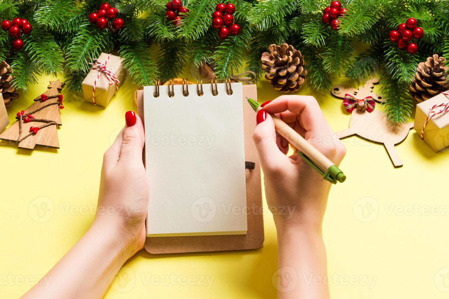 Top view of female hand making some notes in noteebok on yellow background. New Year decorations and toys. Christmas time concept photo