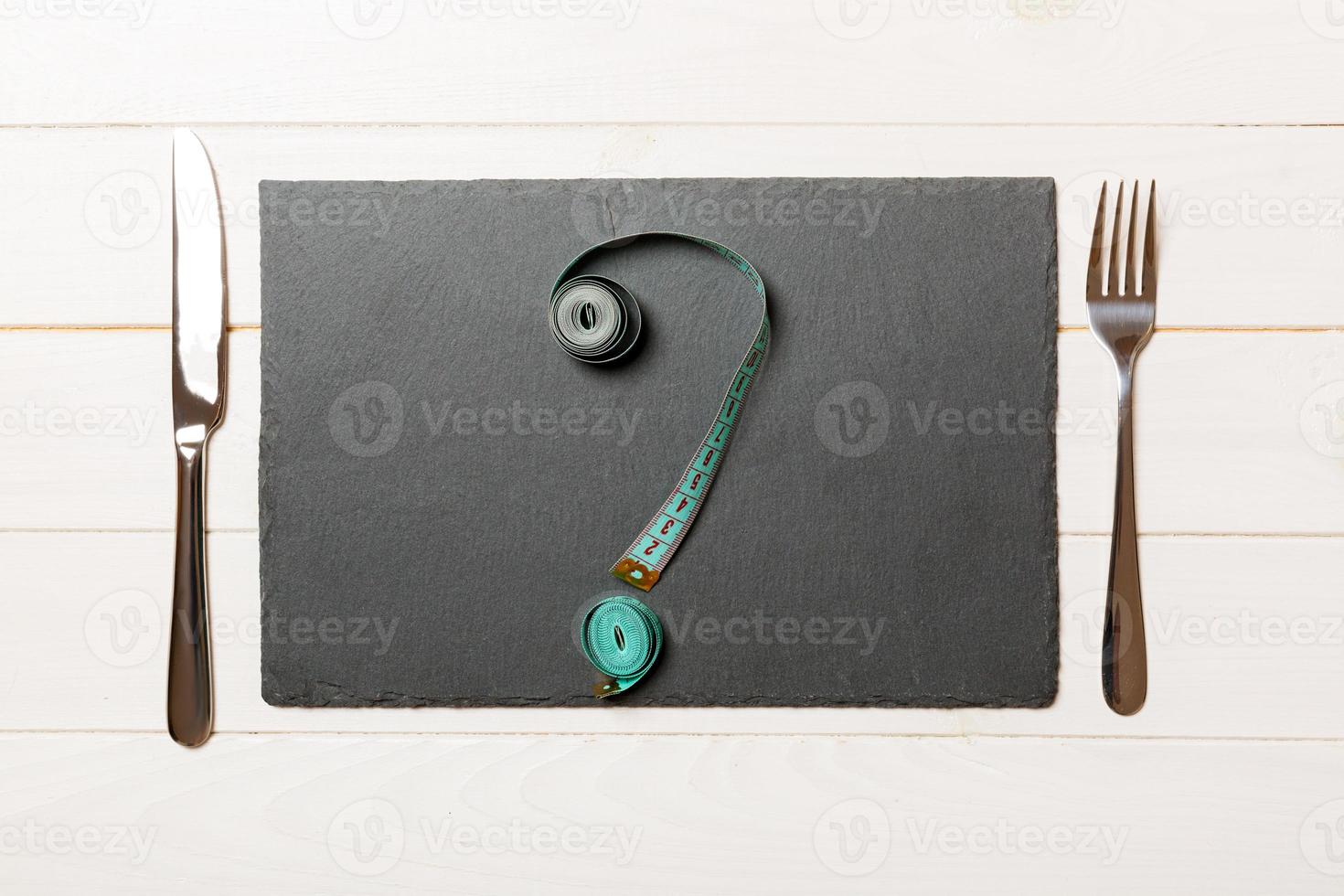 Top view of slate plate, fork and measure tape in form of question mark on wooden background. Overeating concept and copy space photo