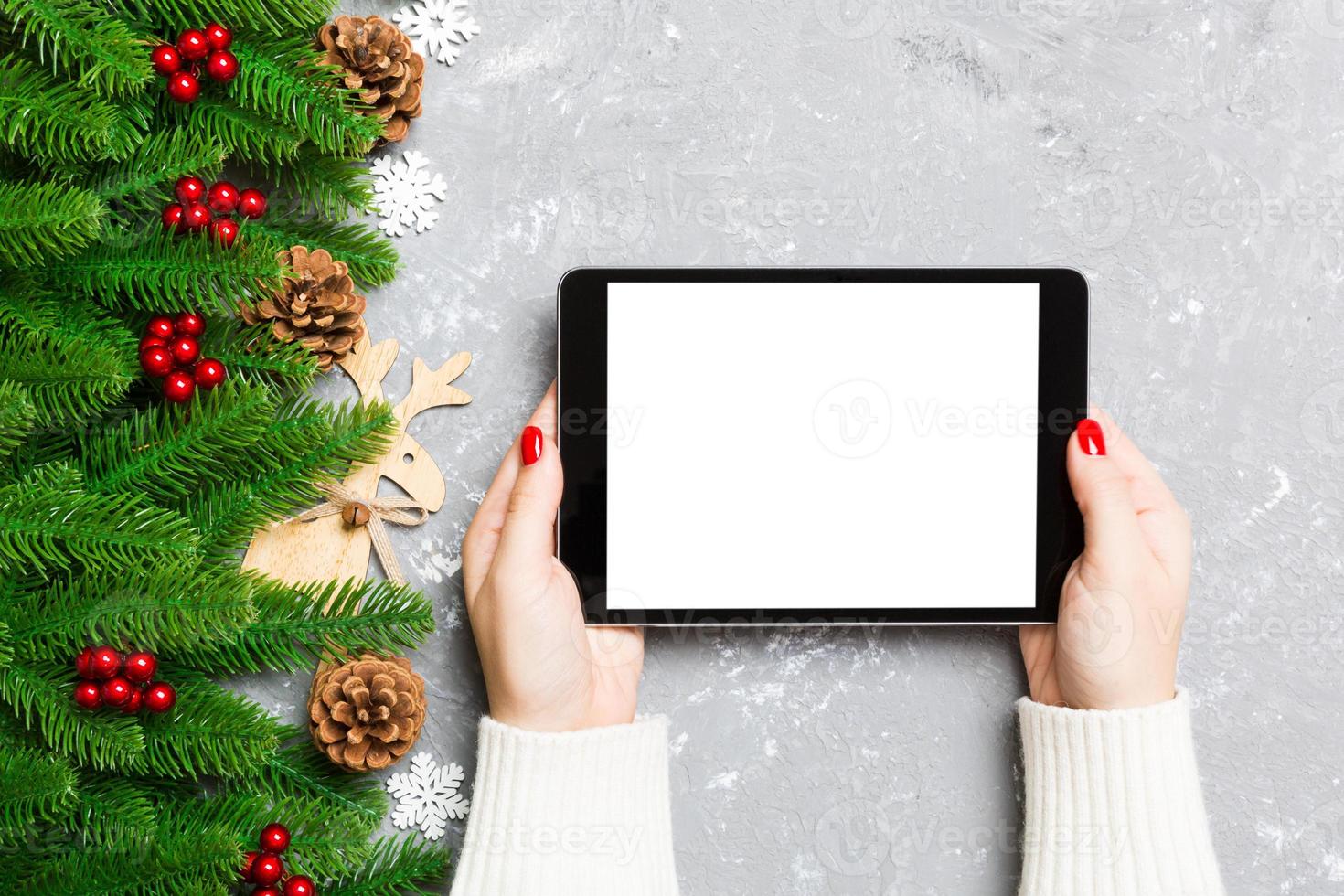 Top view of female hands holding a tablet on cement Christmas background made of fir tree and festive decorations. New year holiday concept. Mockup photo