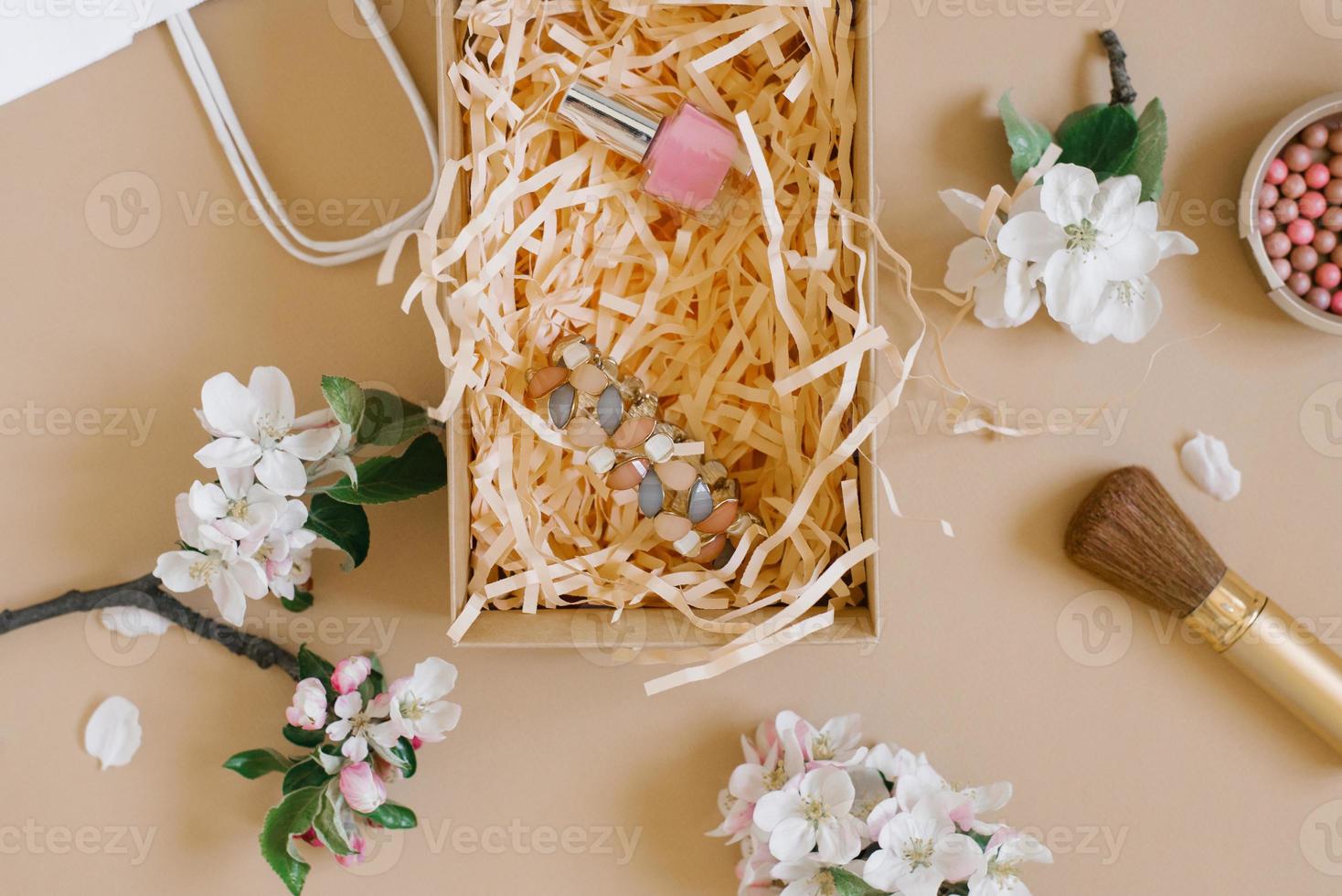 Blush, brush, nail polish and bracelet on a beige background surrounded by white delicate flowers in a gift box. Flat lay photo