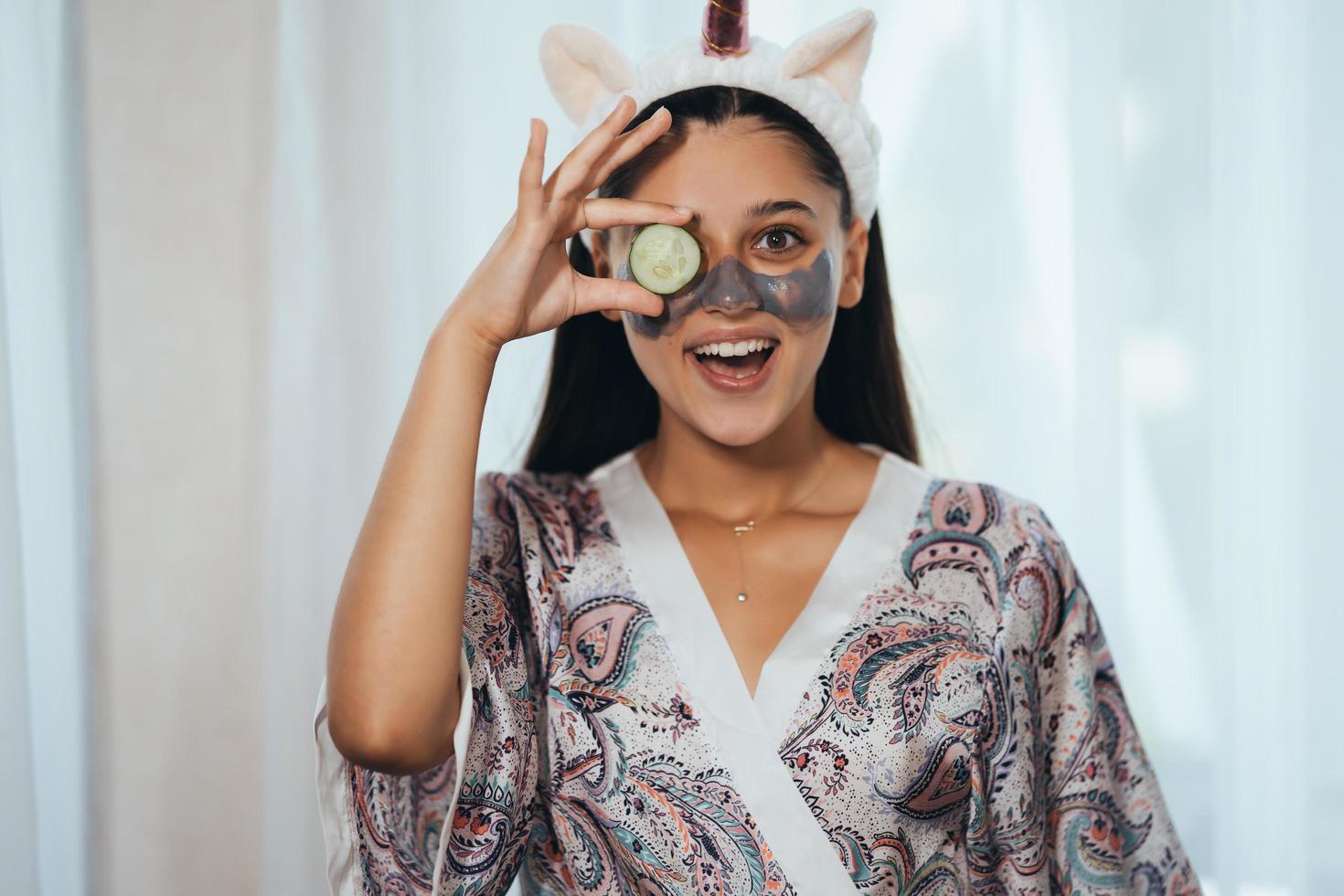 una mujer de spa sonriente y divertida con una máscara facial fresca sostiene pepino. foto