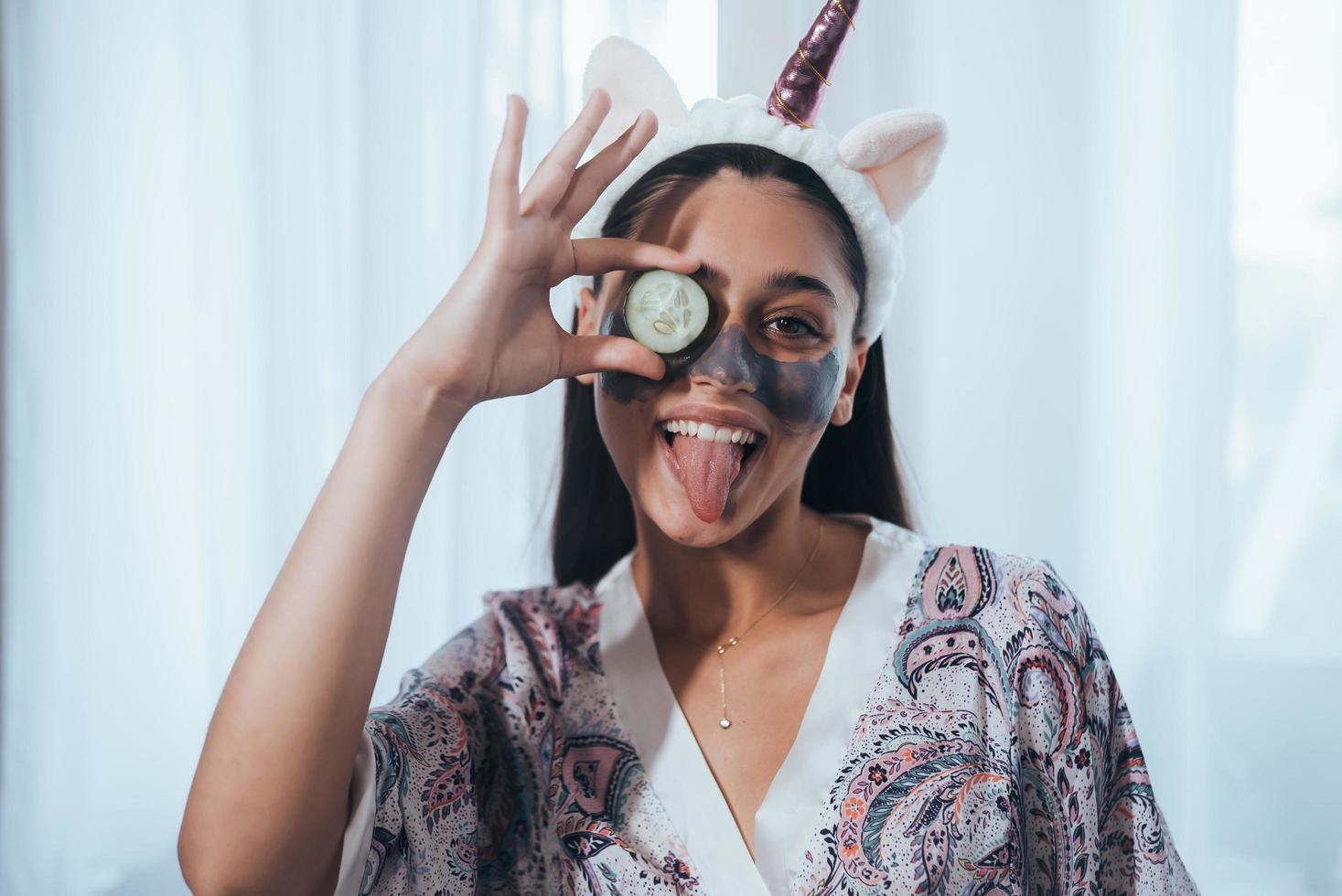 una mujer de spa sonriente y divertida con una máscara facial fresca sostiene pepino. foto