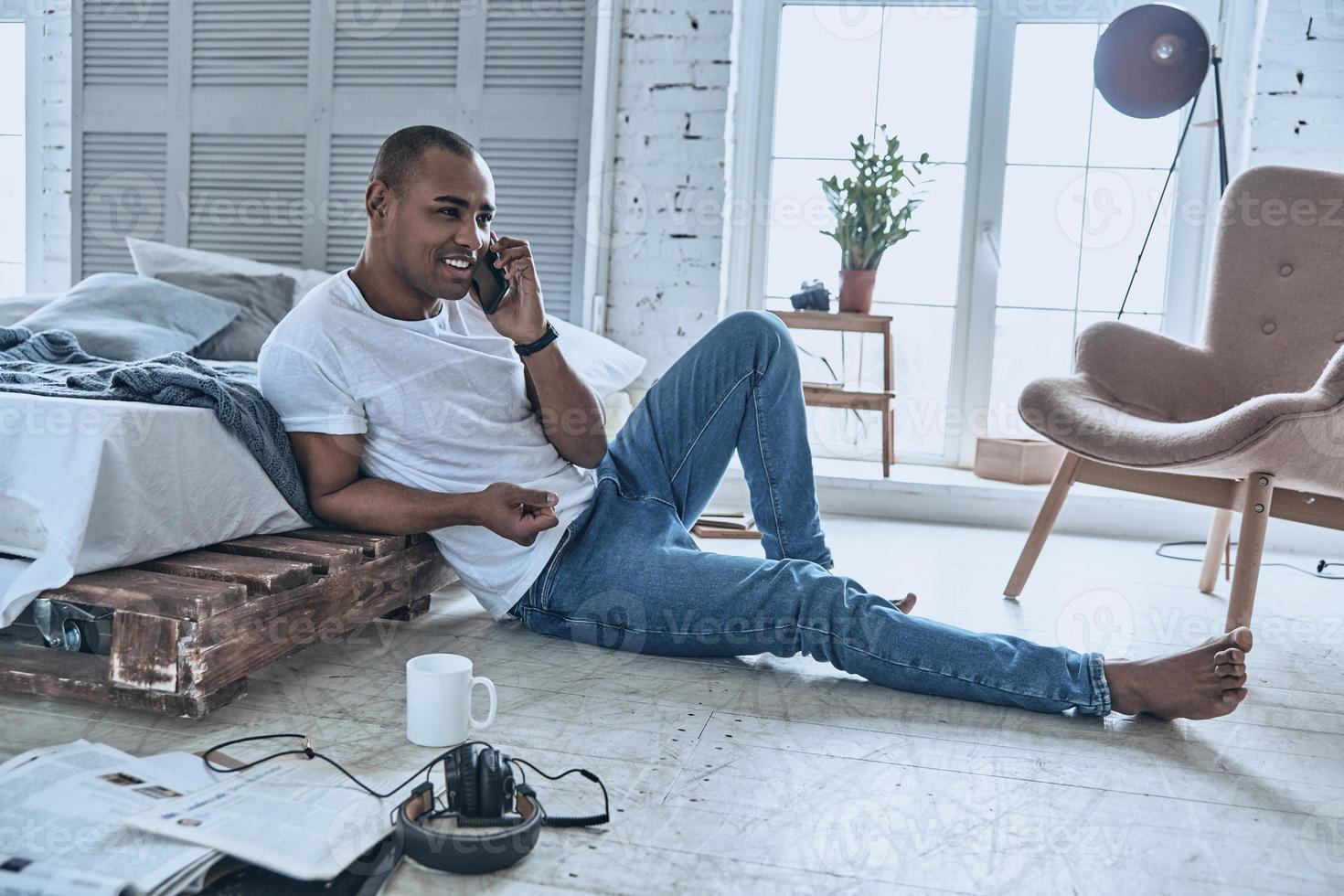 Glad to hear you Handsome young African man talking on the phone and smiling while sitting on the floor at home photo