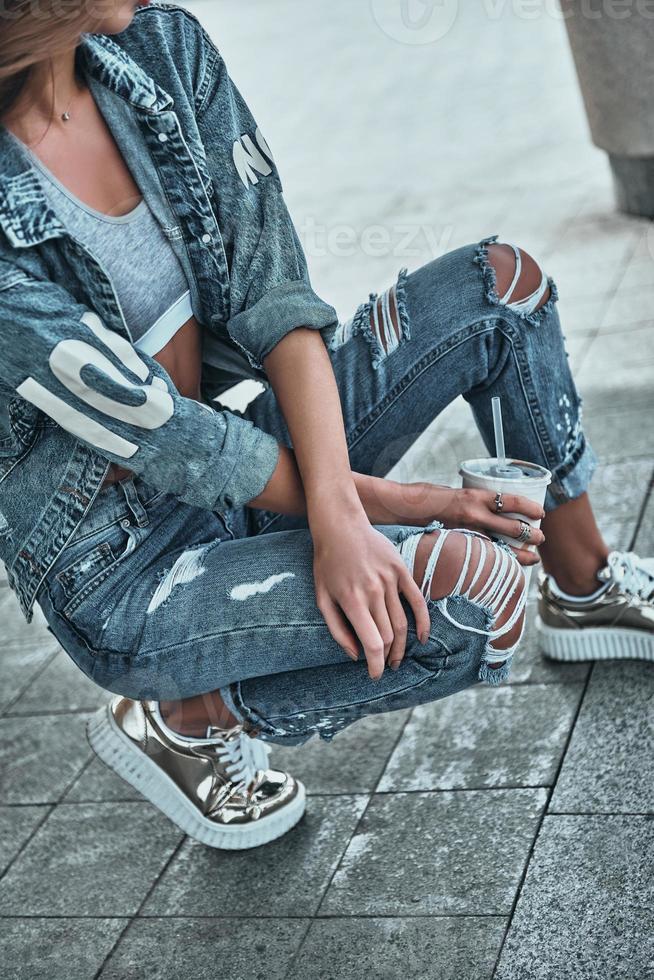 joven y hermoso. primer plano de mujer joven en jeans usar ropa casual sosteniendo una taza desechable mientras se agacha al aire libre foto