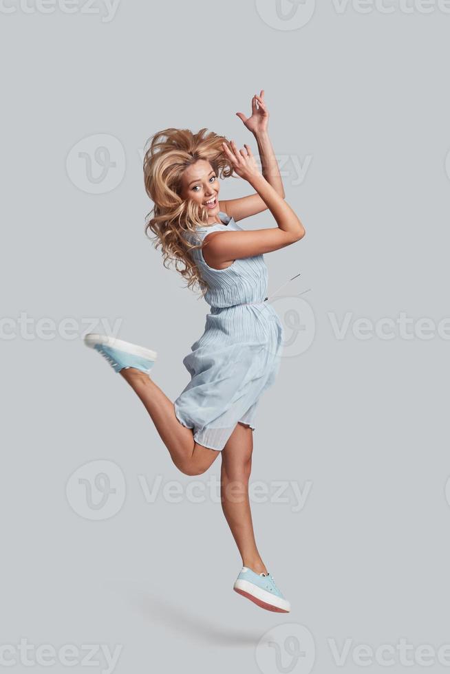 Full length studio shot of playful young woman looking at camera and smiling while jumping against grey background photo