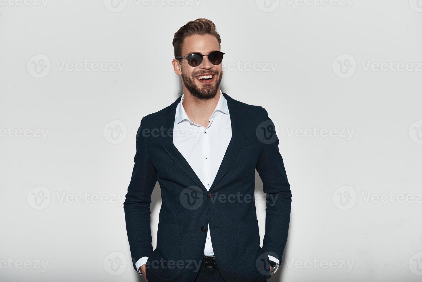 Looking just perfect. Handsome young man in full suit keeping hands in pockets and looking away with smile while standing against grey background photo