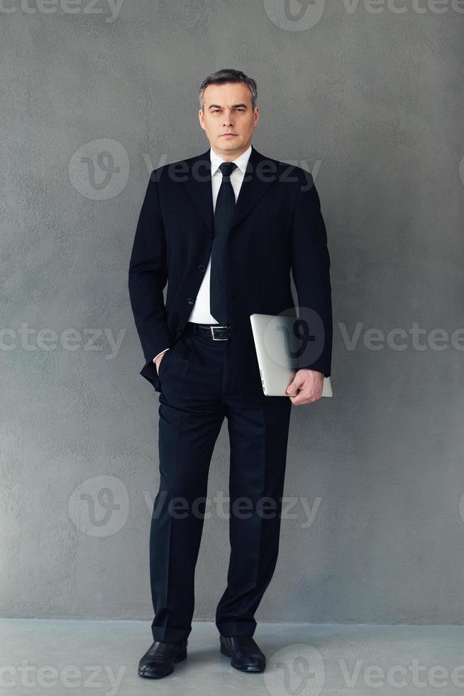Confidence with technologies. Full length of mature businessman holding laptop and looking at camera while standing against grey background photo