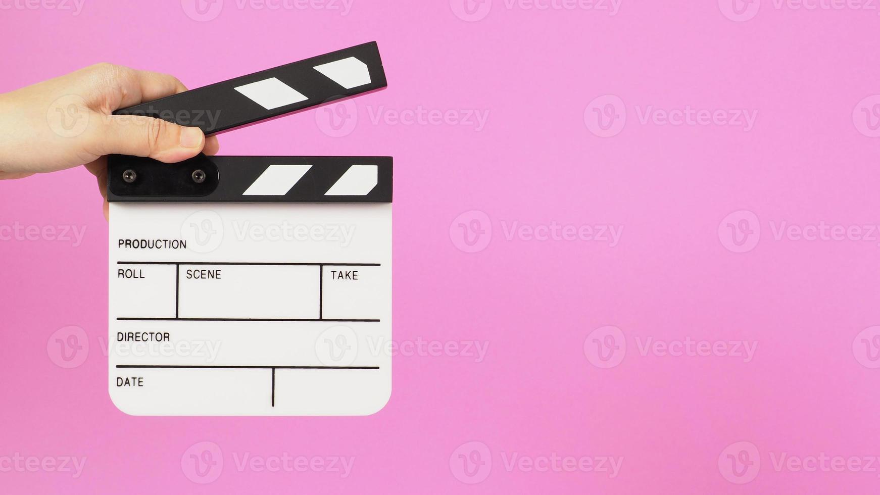 One hand is holding a small white clapper board or movie slate on pink background. side view photo