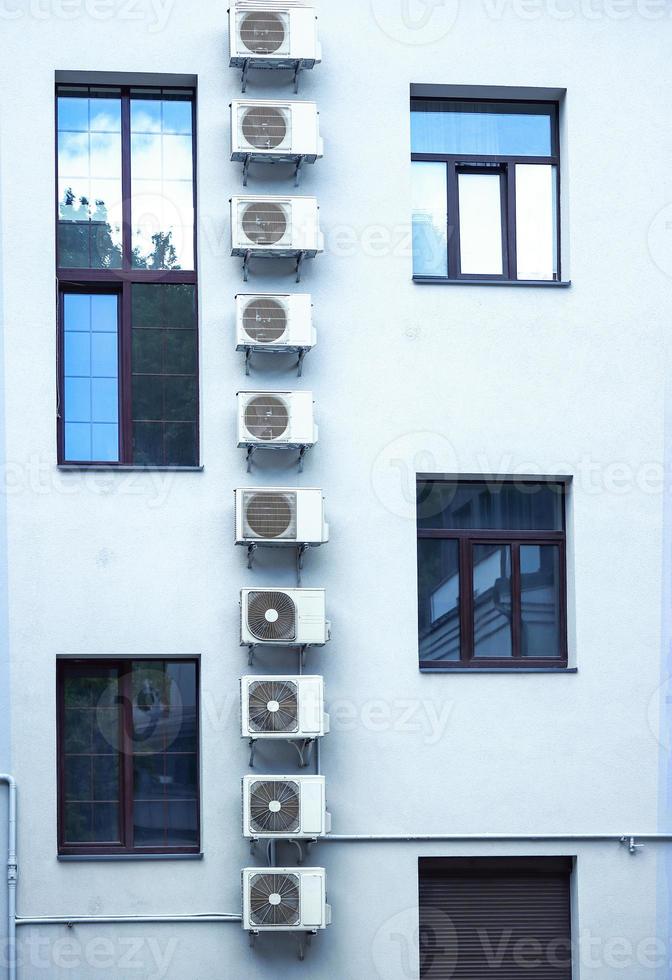 Vertical row of air condicioners on white building with asymmetrical windows with blue sky reflection in windows glass photo